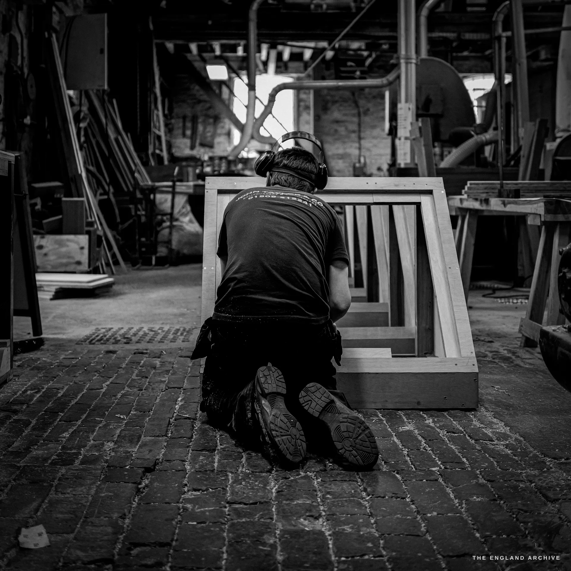 A worker in ear defenders kneeling on the brick floor working on a tall wooden mould frame, seen from behind, the workshop receding behind him.