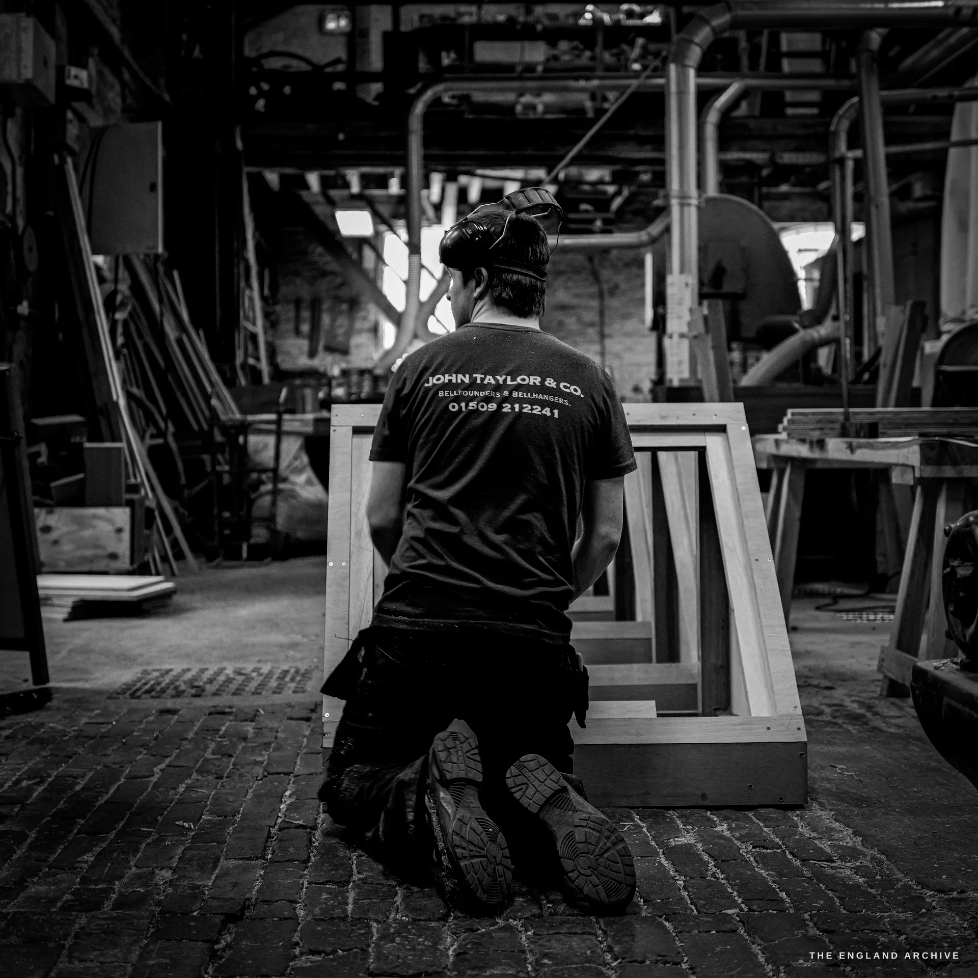 A worker kneeling on the brick floor seen from behind, assembling a wooden mould box, the company name and phone number across the back of his t-shirt.