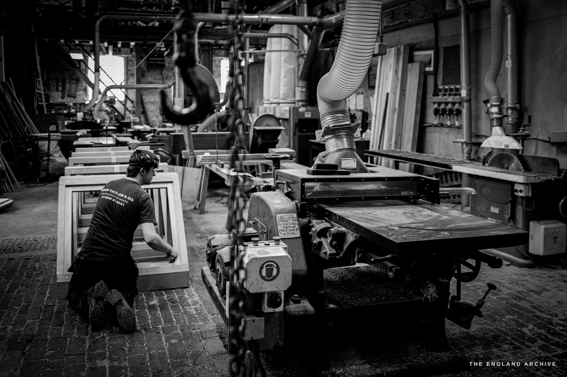 A worker in a John Taylor & Co t-shirt kneeling at wooden mould frames beside a circular-saw bench and extraction ducting, a hoist hook and chain in the foreground.