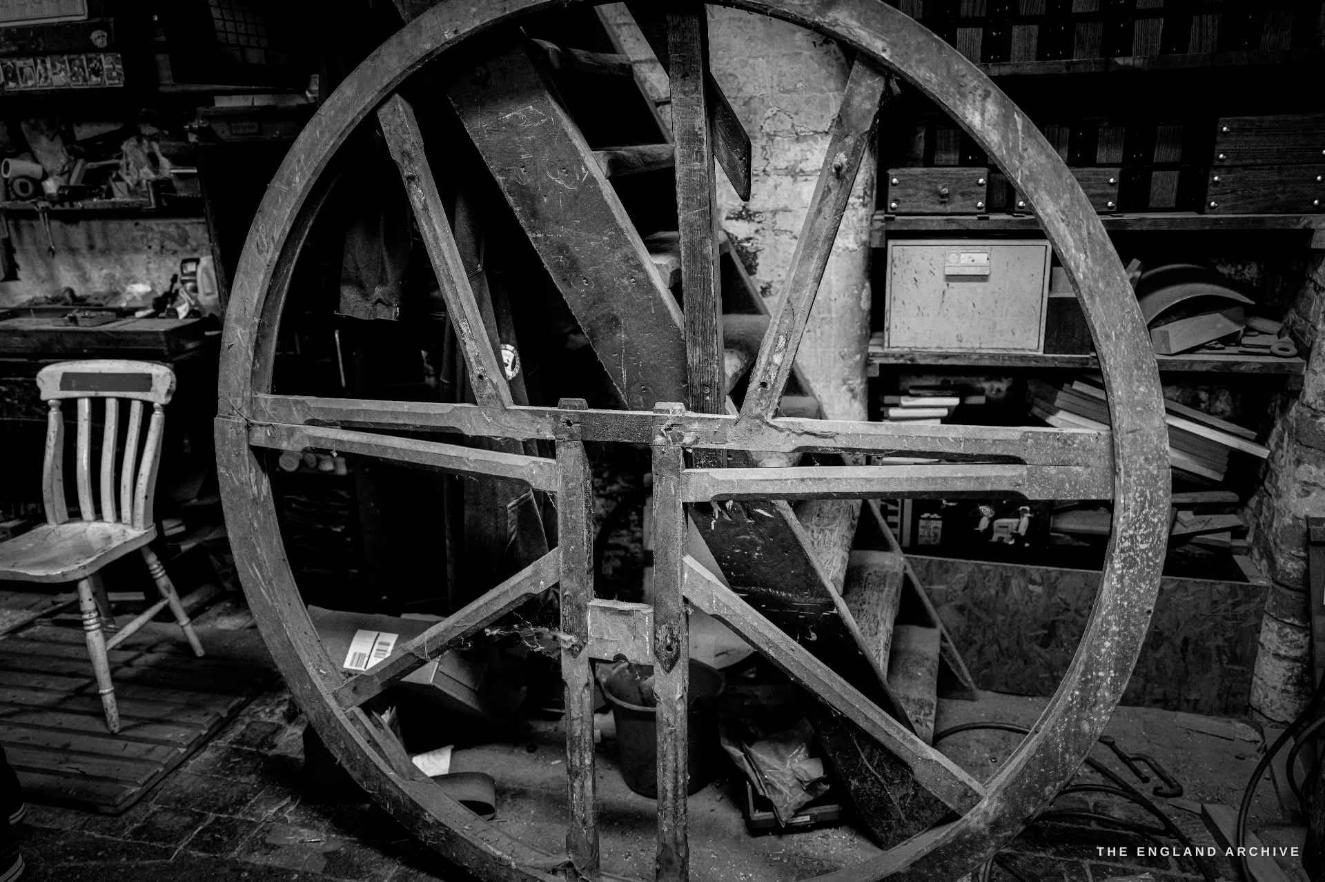 A large spoked wooden bell wheel leaning in a cluttered corner of the workshop, an old wooden chair beside it and banks of drawers behind.