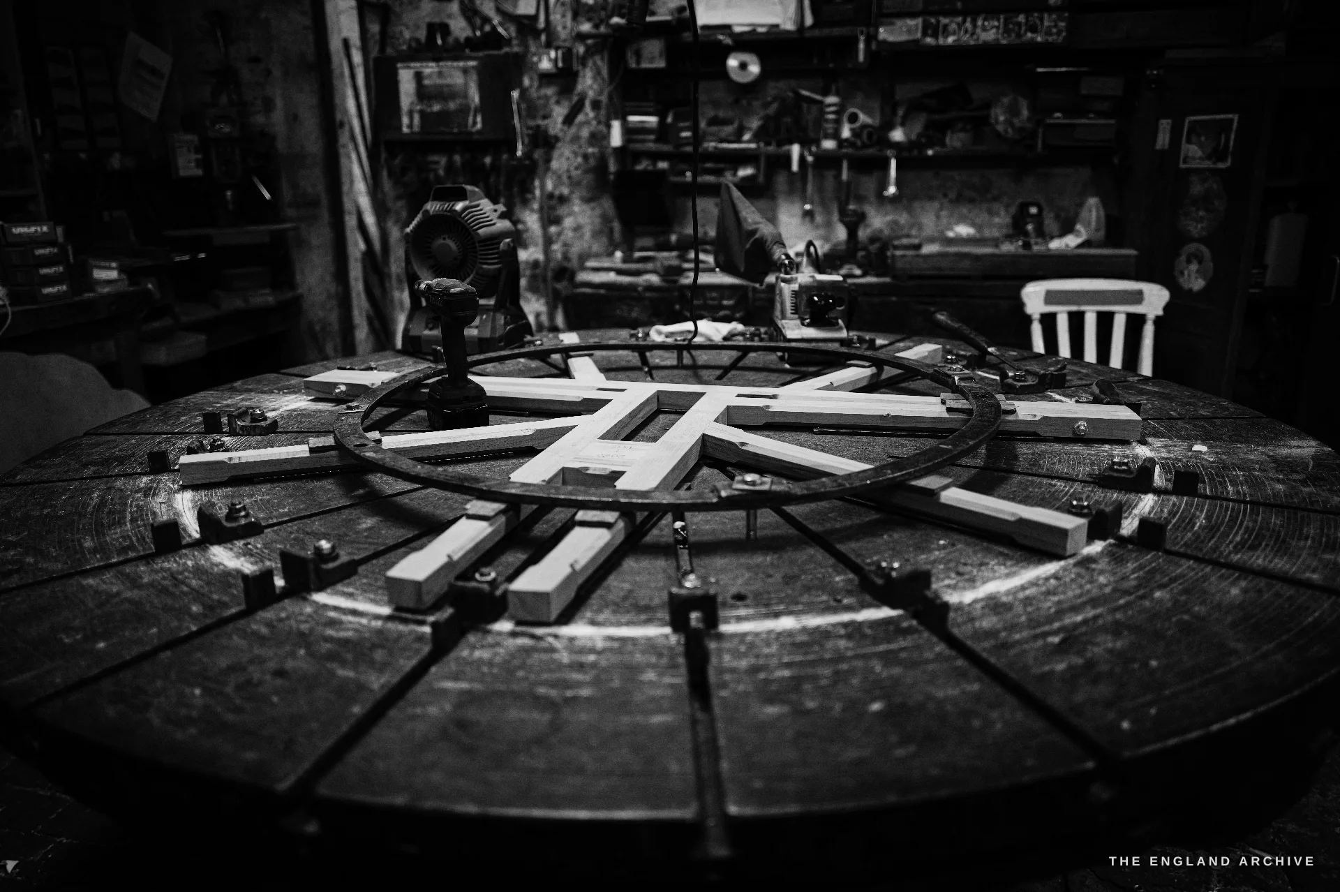 A large circular wooden moulding turntable with a radial arrangement of pattern strips, in the dim pattern workshop, a chair and tool racks behind.
