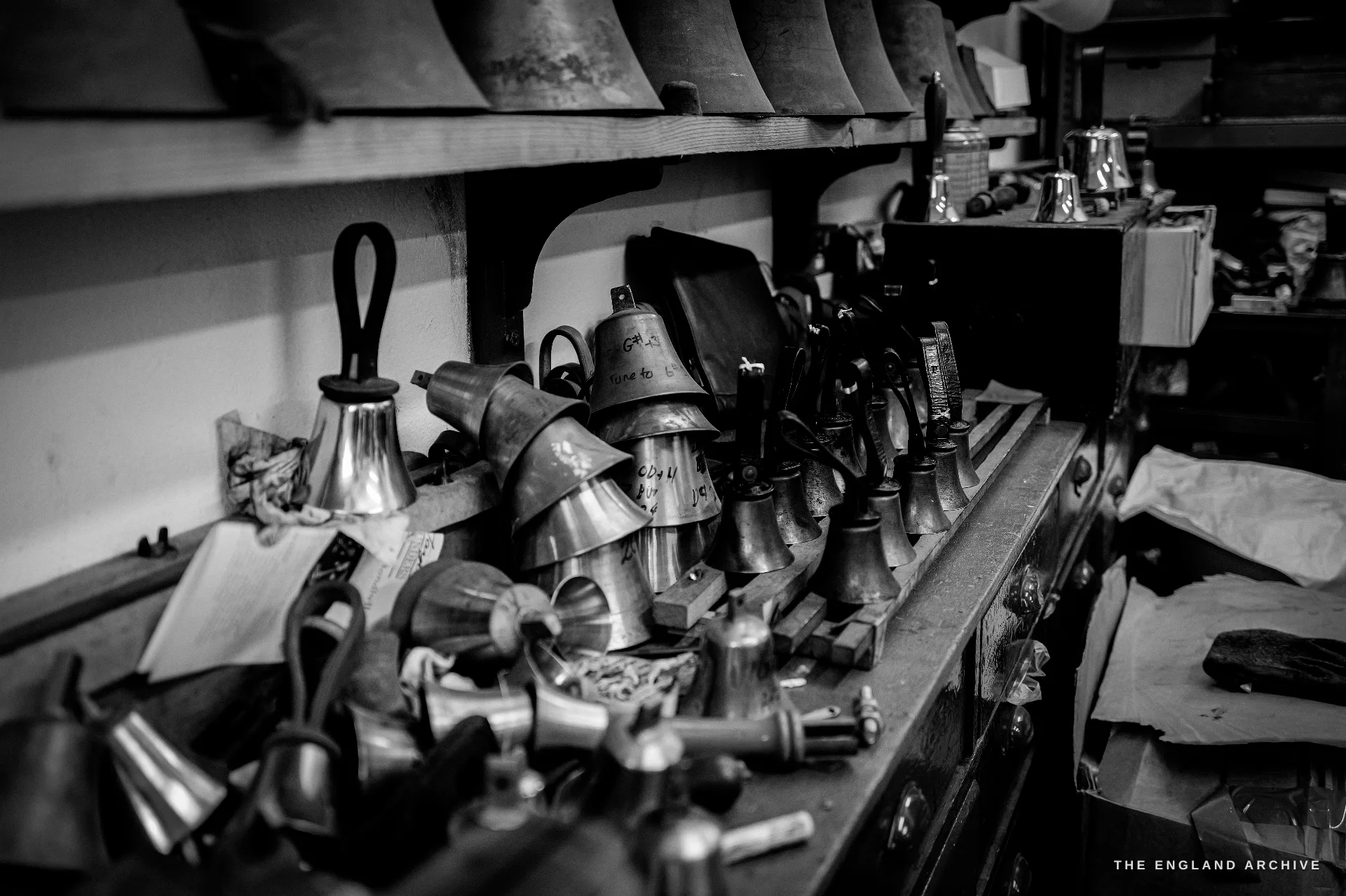 A cluttered handbell-shop workbench crowded with small handbells in various finishes and hand tools, shelves of more bells above.