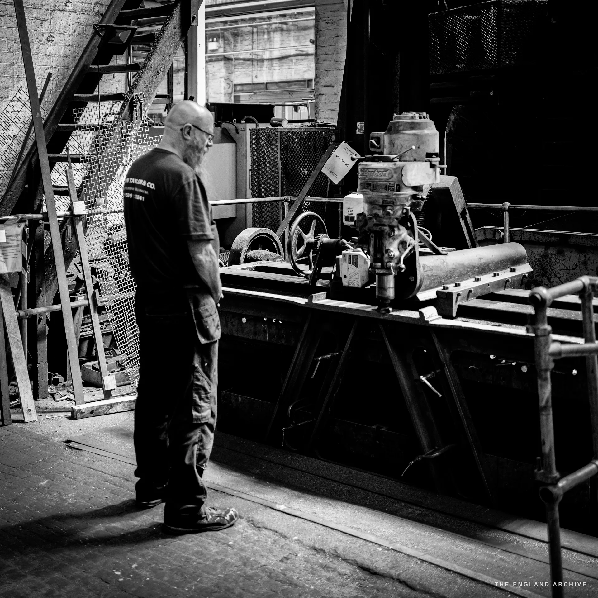 A worker in a John Taylor & Co t-shirt standing at a belt-driven machine bench, seen mostly from behind, an iron stair behind him.
