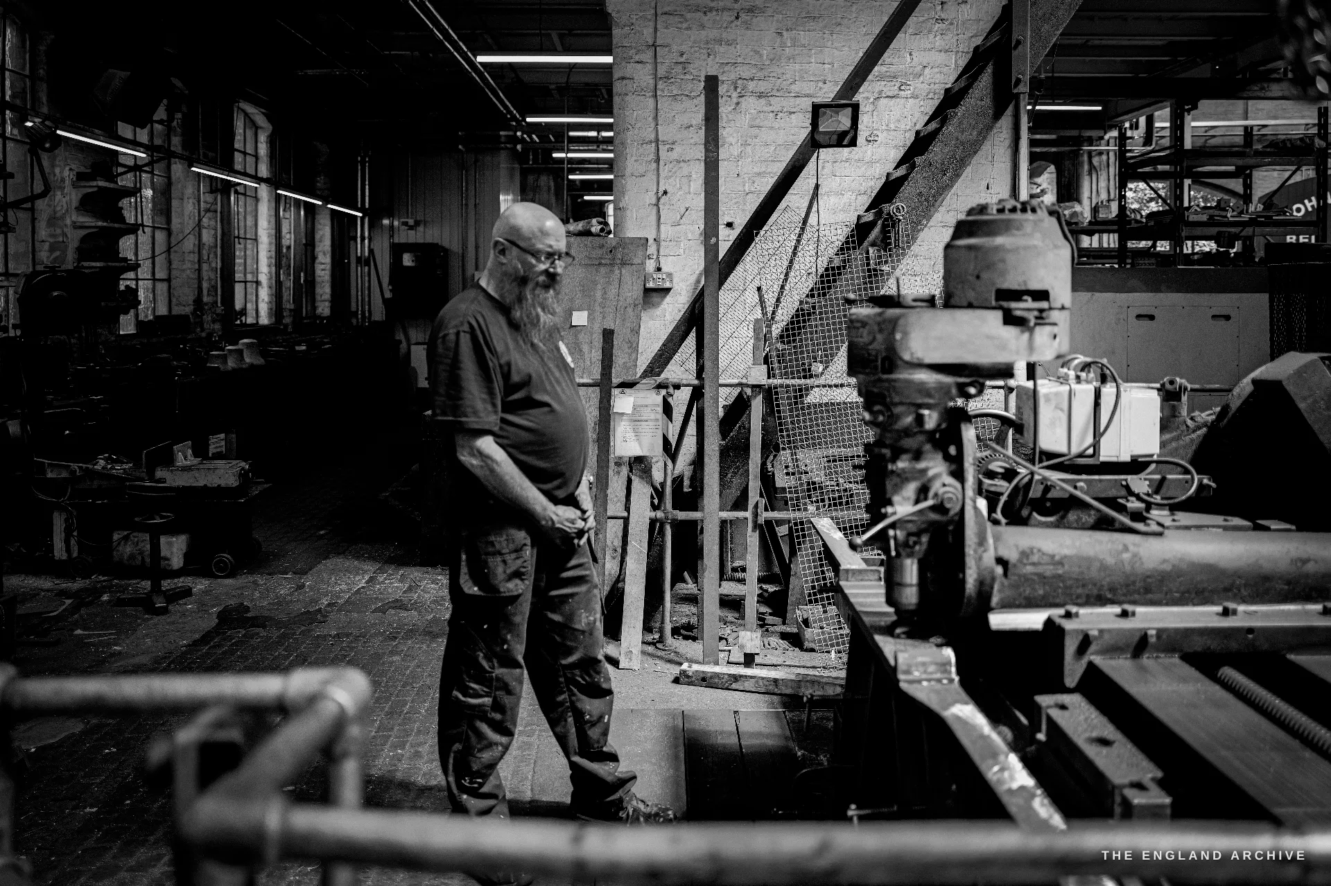 A grey-bearded worker in glasses standing in profile in the workshop, hands clasped, beside heavy machinery, the long hall behind him.