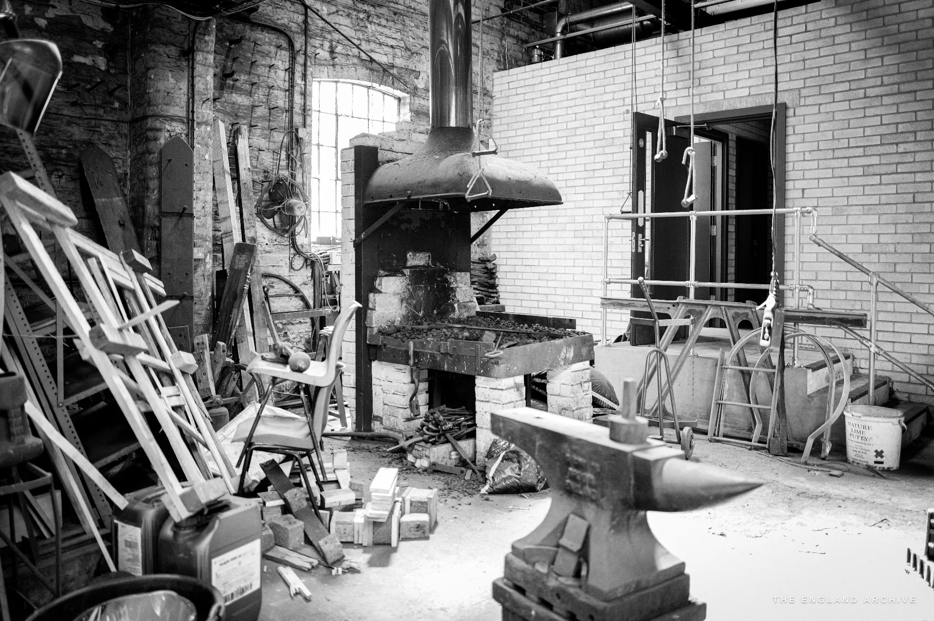 The forge corner of the works: an anvil on a stump in the foreground, a brick hearth with an extraction hood behind, ladders and bell stays stacked, an arched window.
