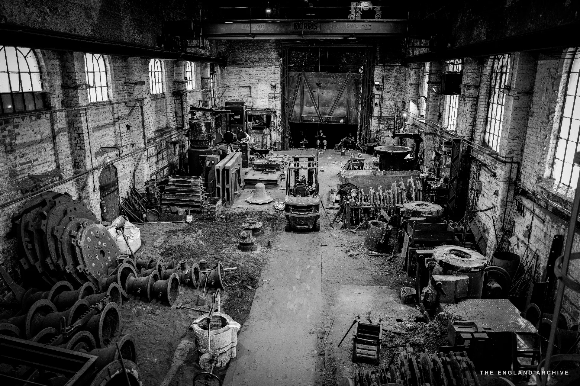 A high wide overview of the moulding and casting shop - sand floor, moulds and ladles, a forklift, an overhead gantry crane, stacked bell wheels at left and tall arched windows.