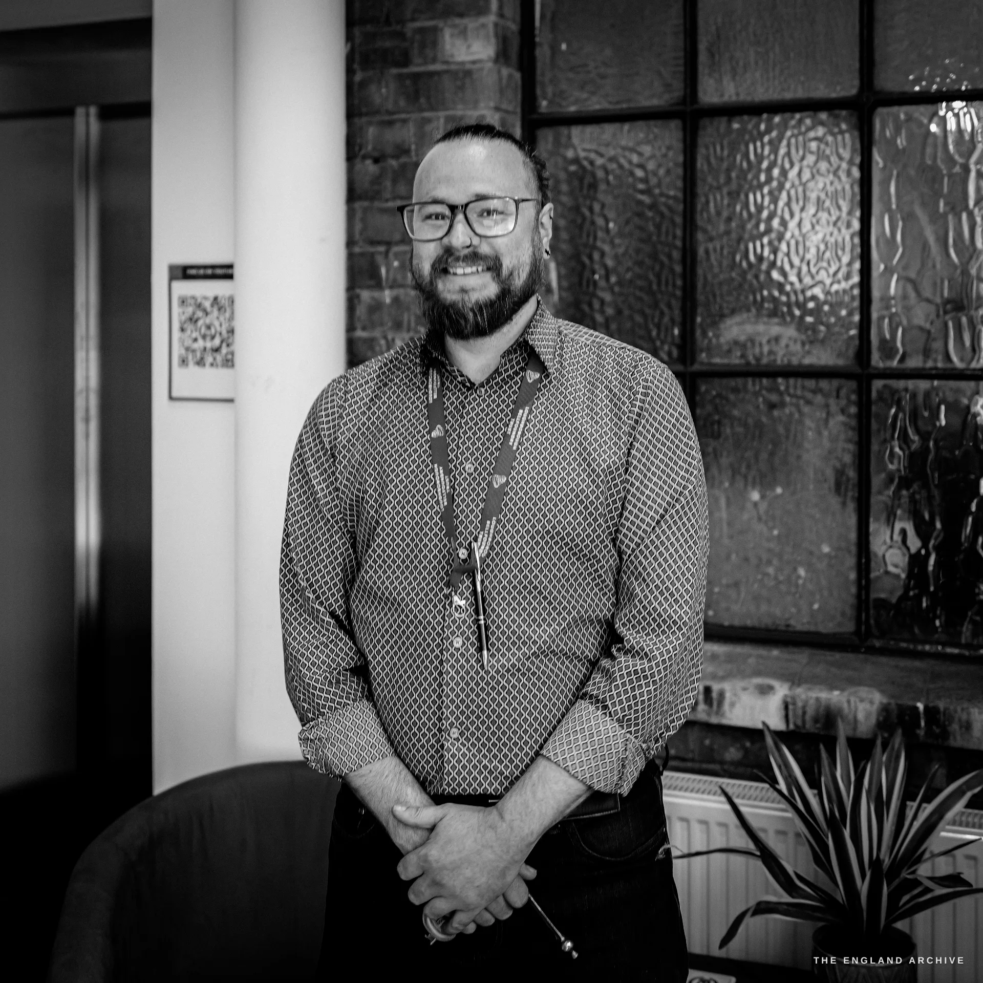 A bearded man in glasses, a patterned shirt and a lanyard standing by a leaded window with a plant, smiling at the camera.