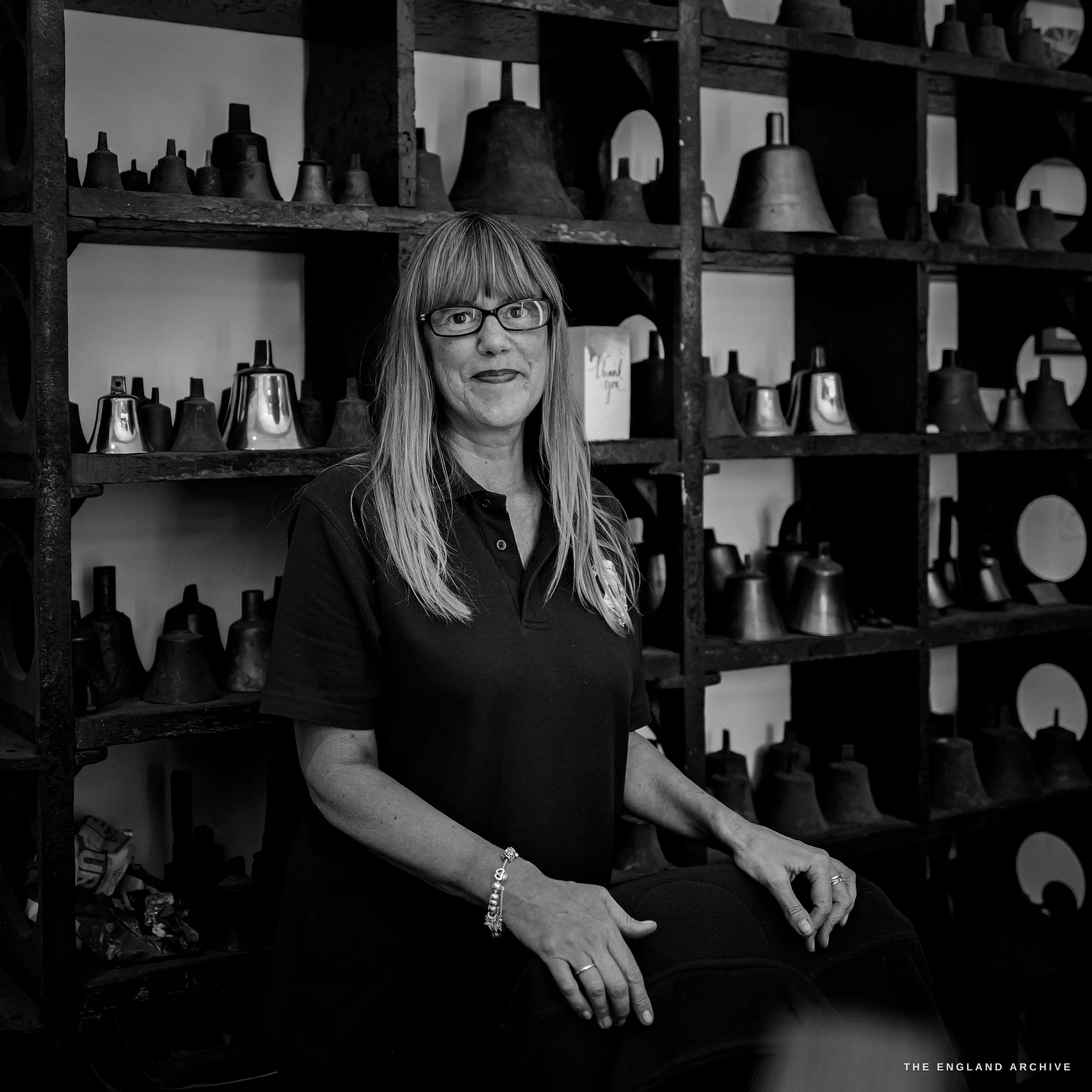 A reception team member with long fair hair and glasses, in a dark polo shirt, seated in front of the shelves of bells at the front desk.