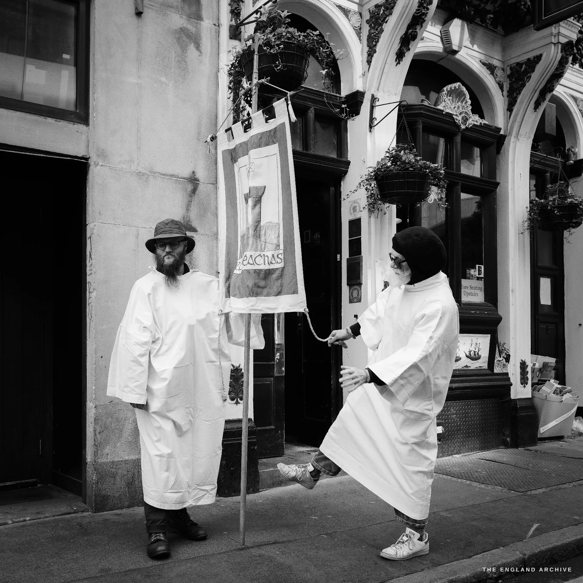 Two members of the Druid Order in white robes outside a Wetherspoon’s pub frontage, one in a dark hat holding a banner pole, the other passing a tasselled cord across the banner of the order.