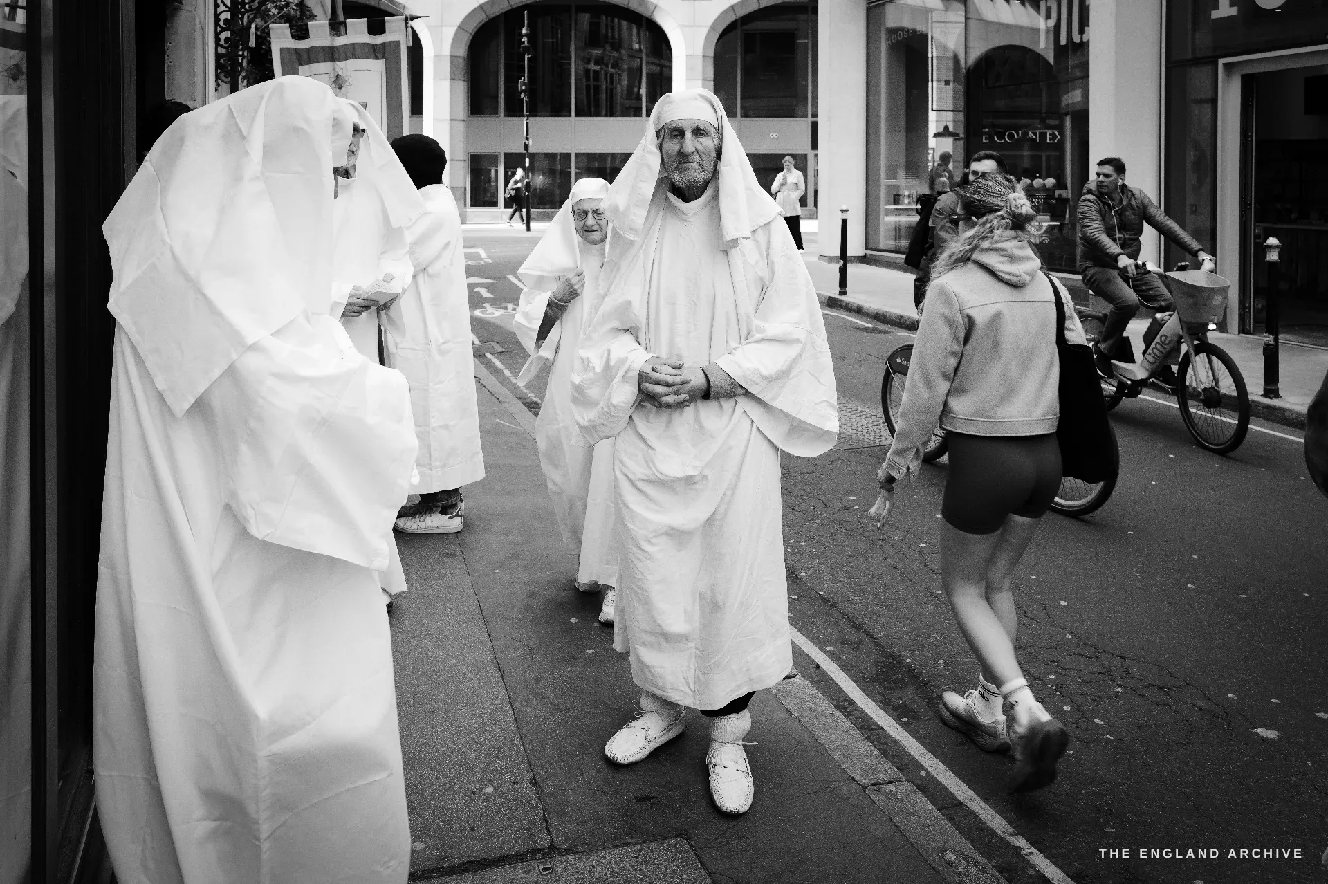 A bearded male Druid in white robes standing in the middle of a City pavement, hands clasped, a young woman in athletic shorts running past him from the right.