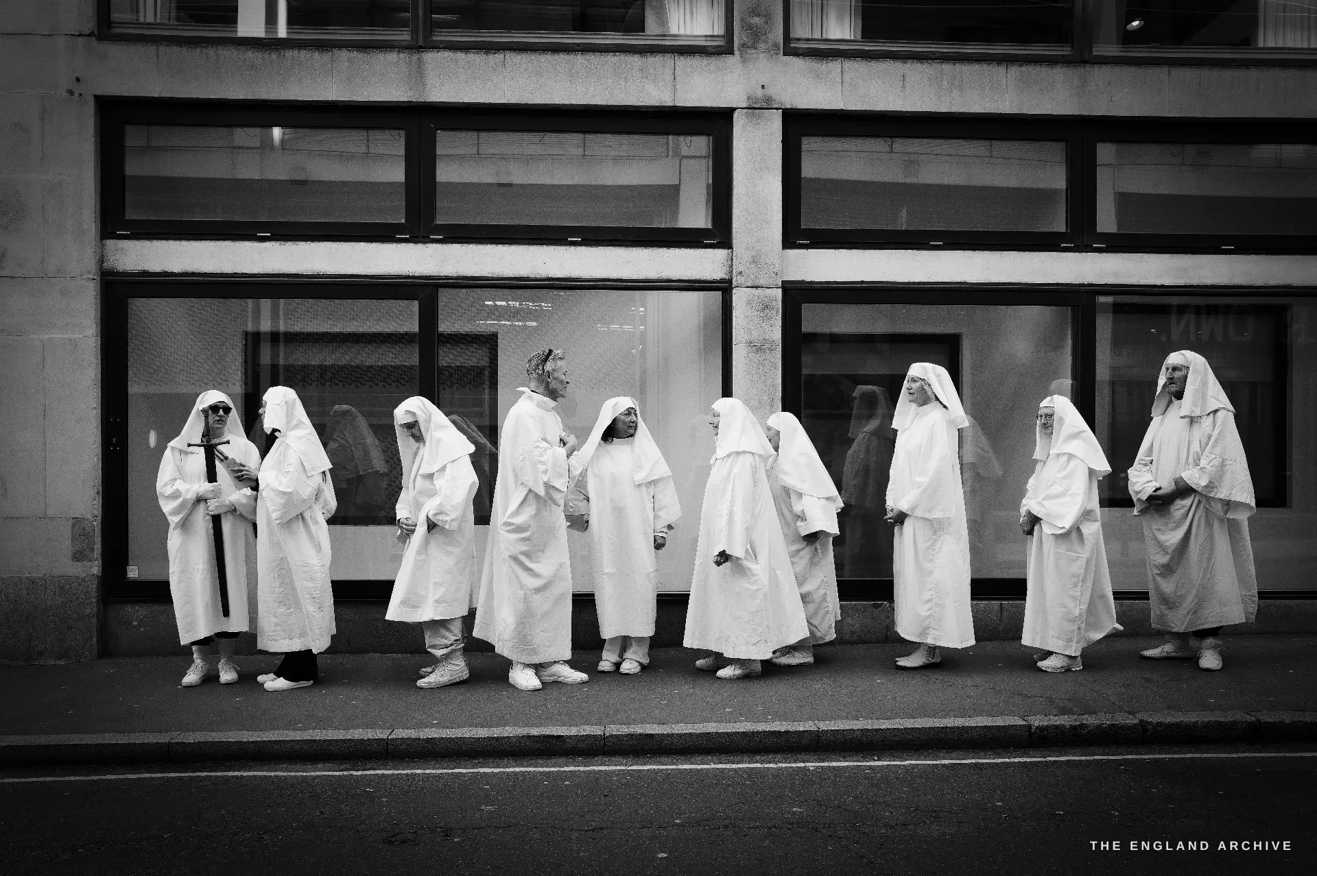 A line of nine or ten white-robed Druid Order members standing along a glass-fronted modern office building, waiting in profile.