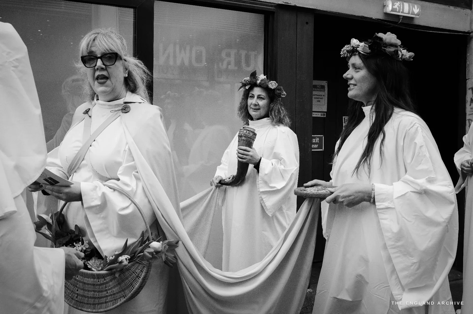 Three women in white ceremonial robes seen close, one with a floral crown holding a stoneware chalice, another beside her wearing a flower wreath in her hair.