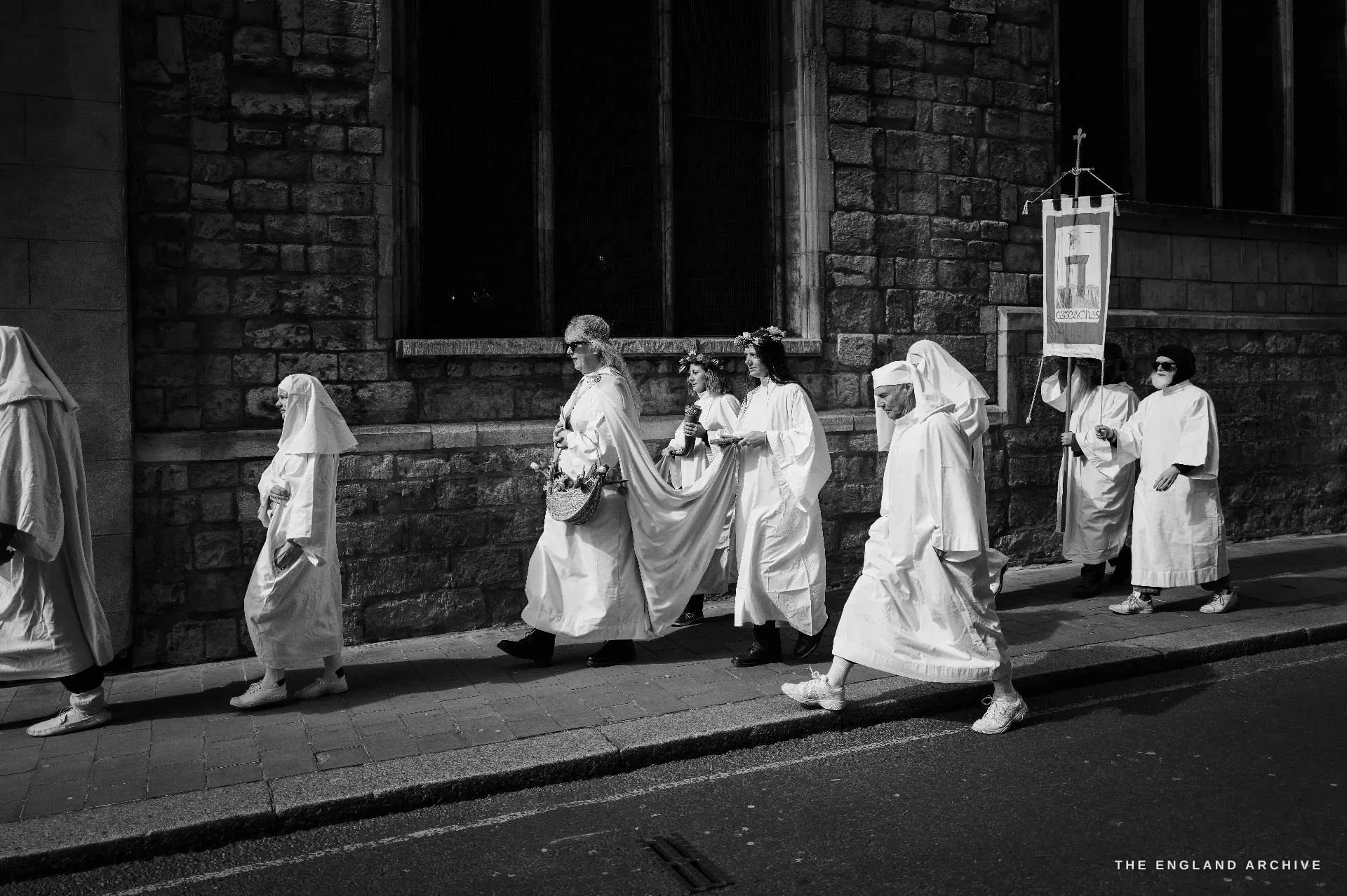 A procession of Druid Order members in white robes walking in single file along a pavement past a stone church wall, the rear of the line carrying an upright embroidered banner of the Order.