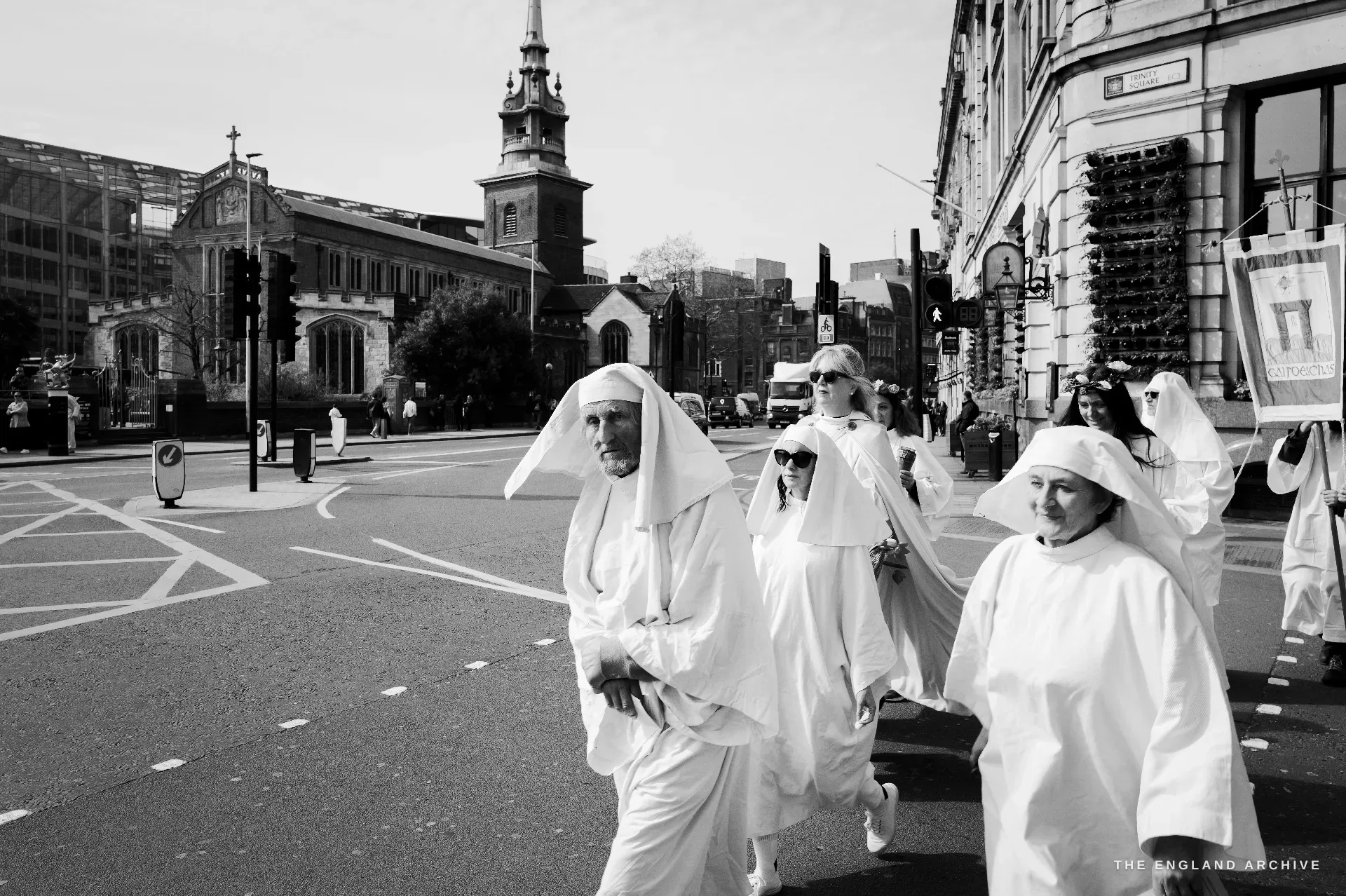 A close formation of Druid Order members crossing a London road, the pale stone tower and white spire of All Hallows-by-the-Tower rising in the background to the left.