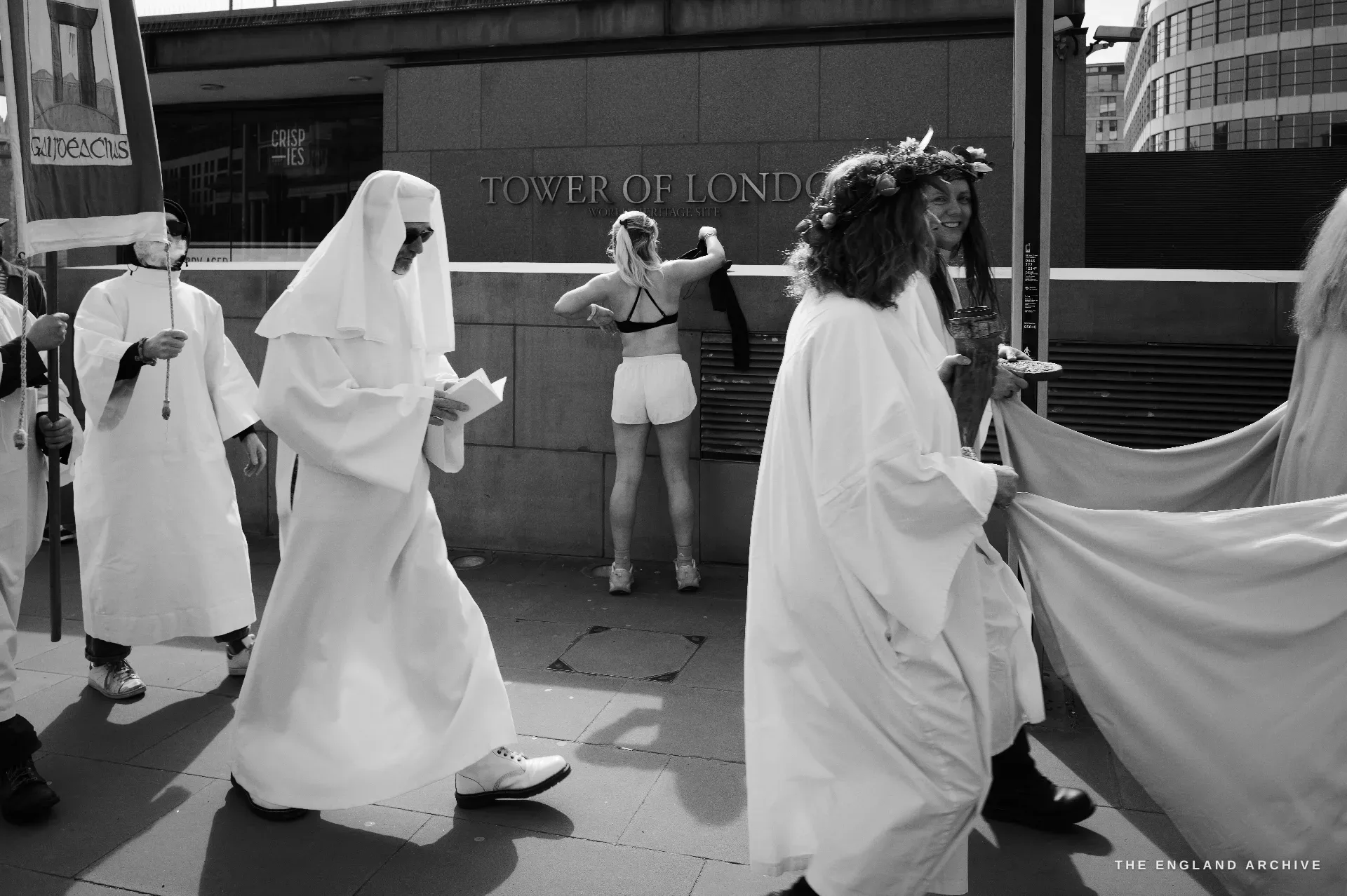 A line of white-robed Druid Order members walking past the carved TOWER OF LONDON sign on the precinct wall, a woman in athletic gear stretching against the wall in the middle of the frame.