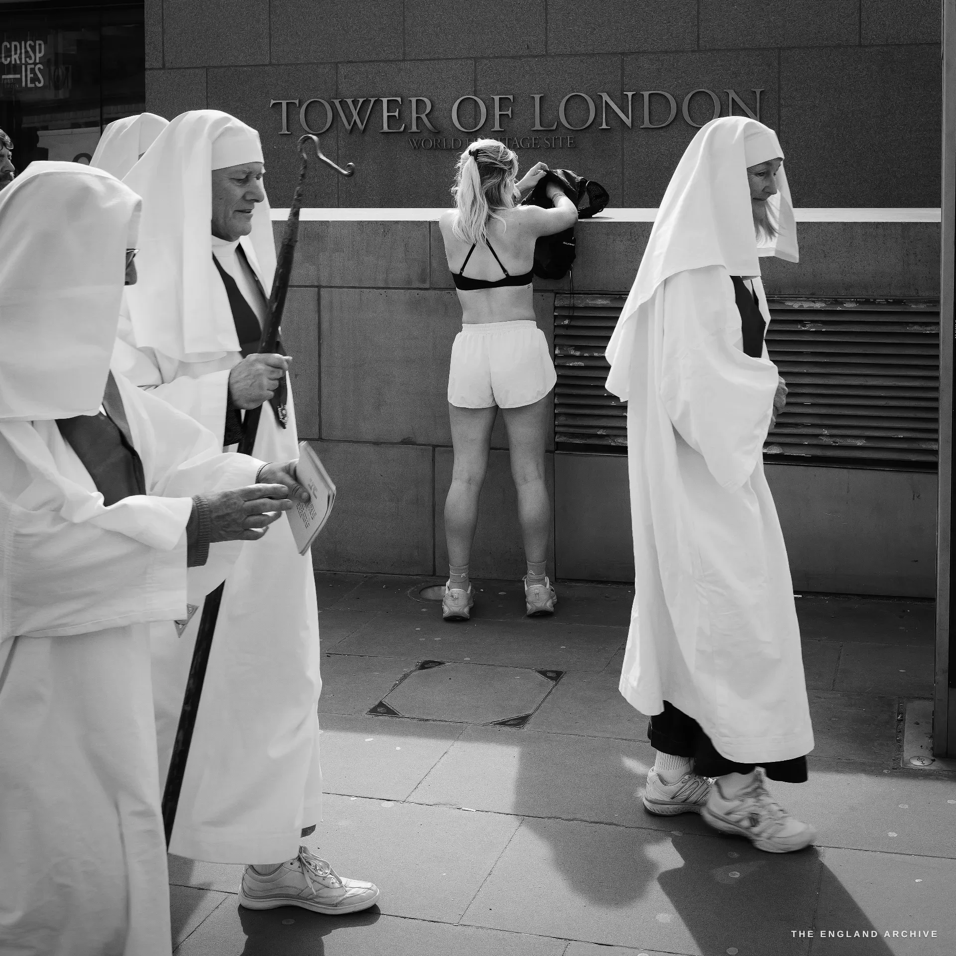 A close view of the carved TOWER OF LONDON inscription on the precinct wall: three Druid Order members in white robes and head-veils in the foreground, a runner in pale shorts stretching against the wall behind them.