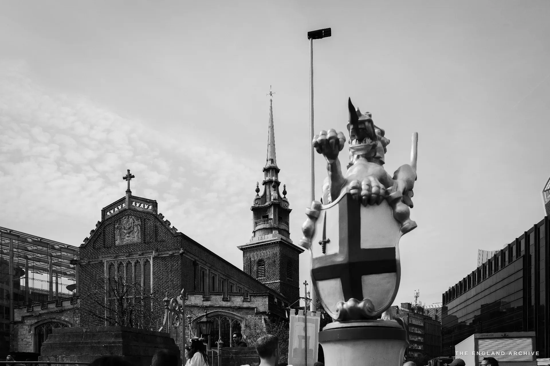 A view across Trinity Square: the brick body of All Hallows-by-the-Tower at left with its small white spire, a sculptural City of London dragon mounted on a pedestal at right against a pale sky.