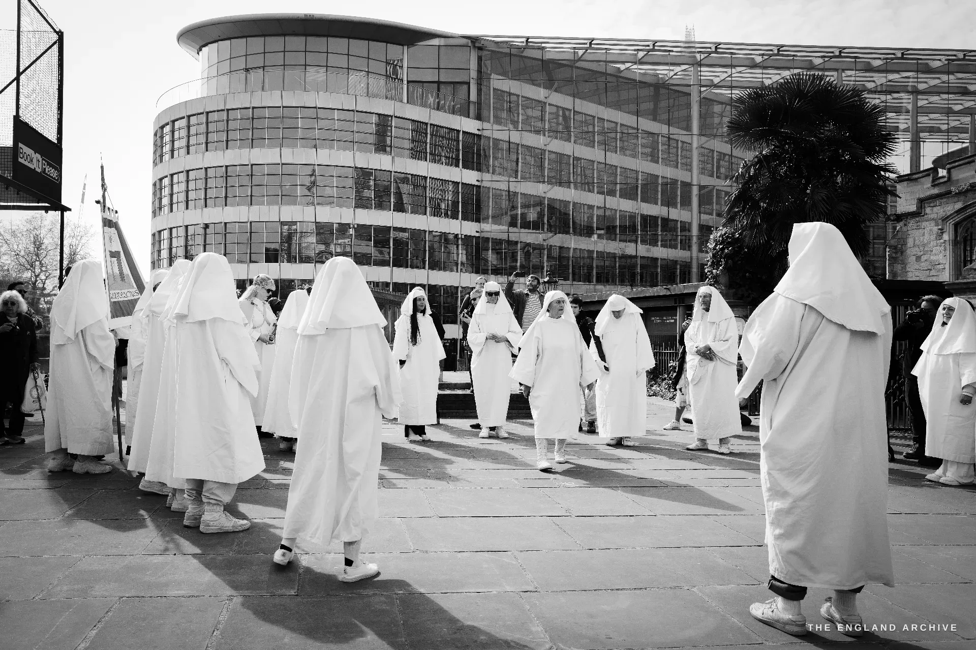 A loose ring of Druid Order members in white robes forming a circle on a paved courtyard in front of a curved modern glass office building, a small palm tree at right.