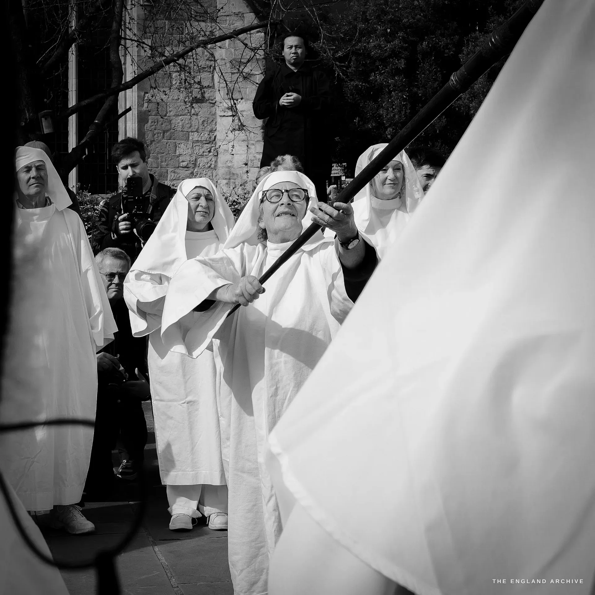 A central Druid in white robes and head-veil raising a tall flag pole to the vertical, surrounded by other Order members and an onlooker in dark clergy dress watching from the rear.