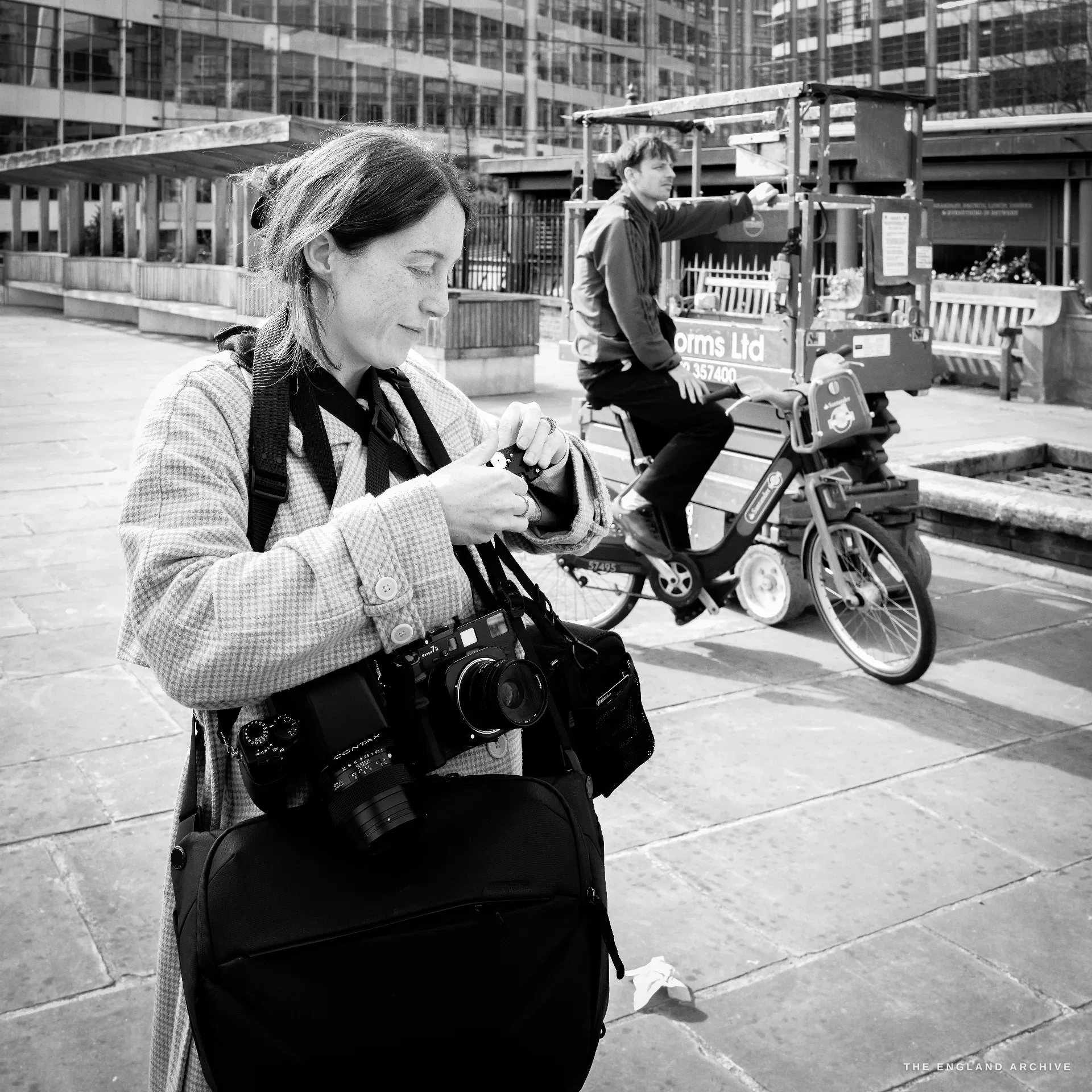 A documentary photographer in a checked coat standing on a London plaza adjusting one of two film cameras hung around her neck, a Santander hire bike and a lift platform in the background.