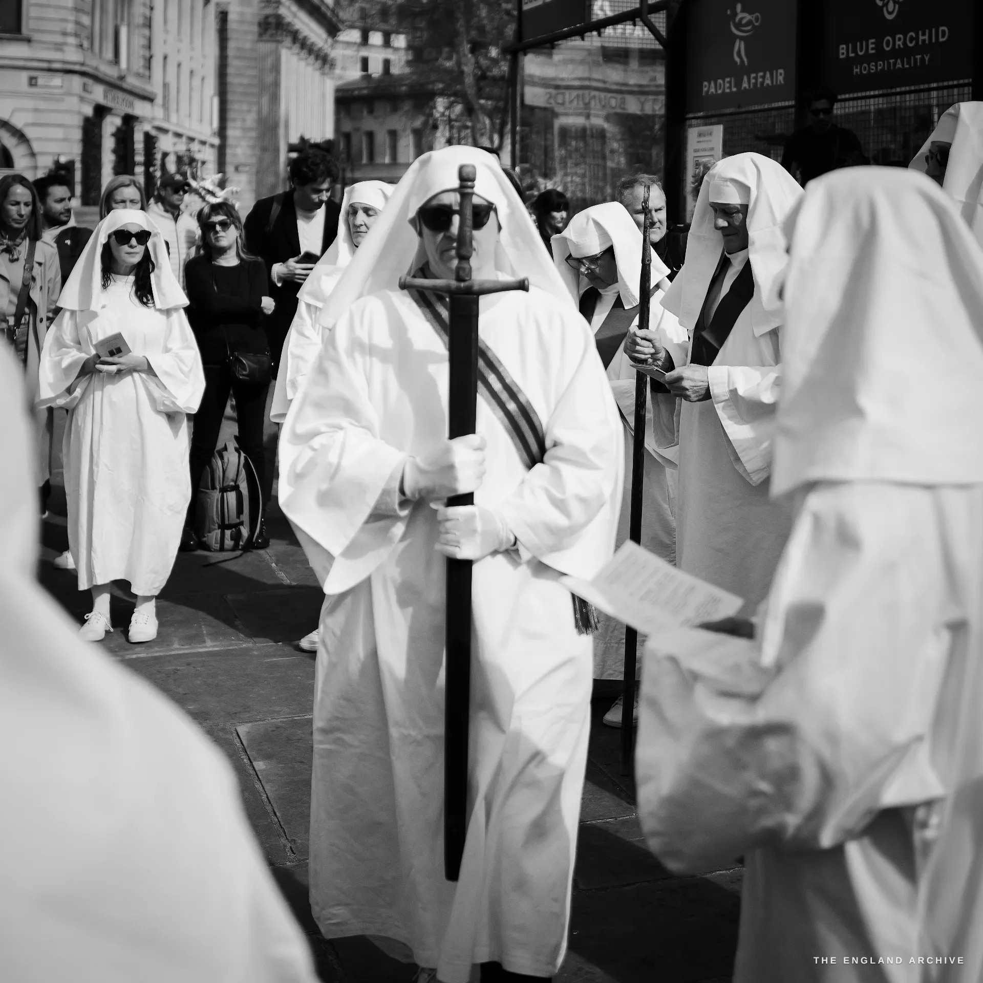 A central robed Druid carrying a vertical sword resting against his shoulder, surrounded by other Order members and an audience of bystanders, the storefront of a hospitality venue visible behind.