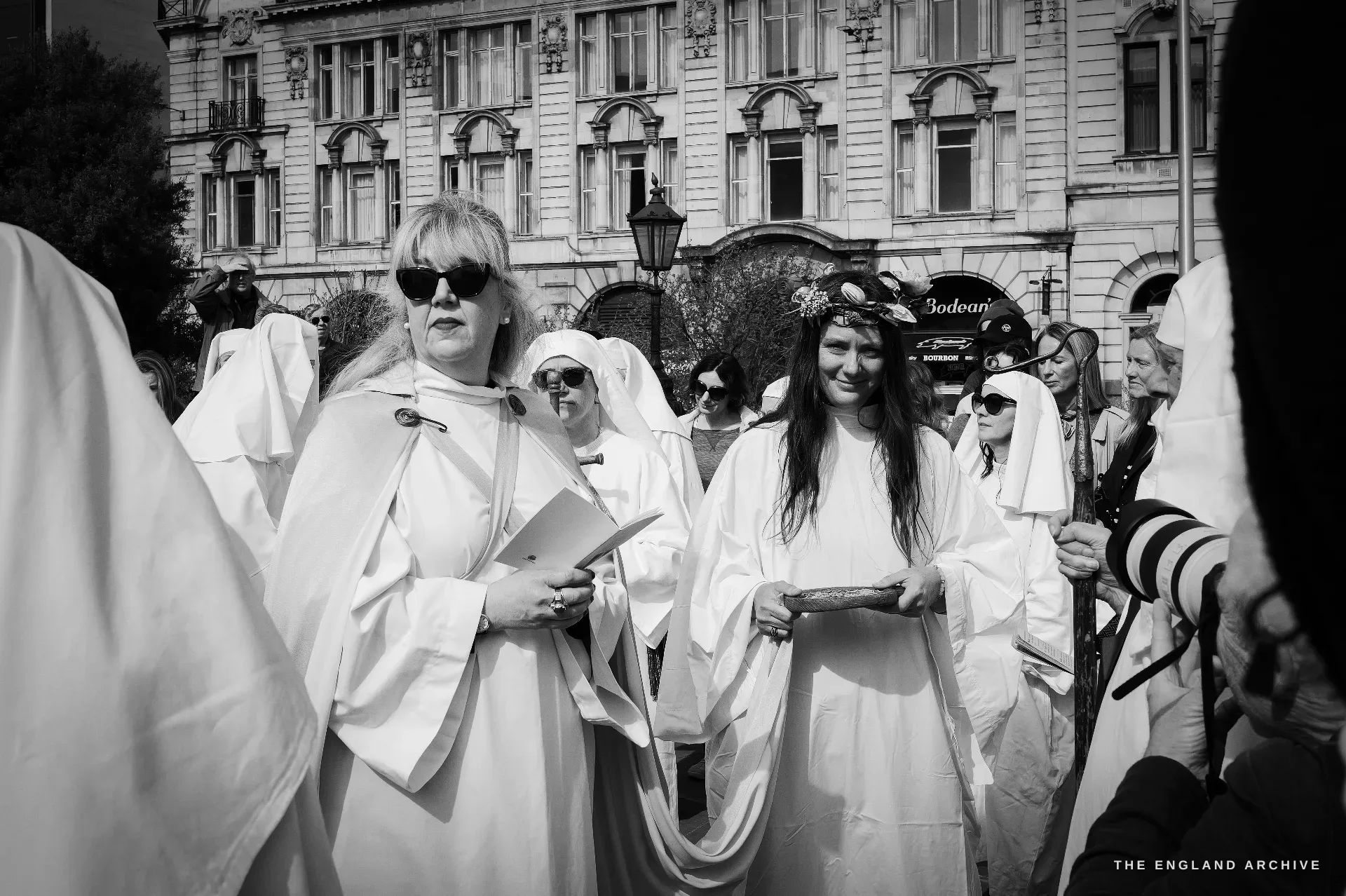 Two female Druid Order members in white robes seen close, both wearing floral crowns - one with sunglasses and a small folder of papers, the other smiling, the white stone facade of a City building behind.