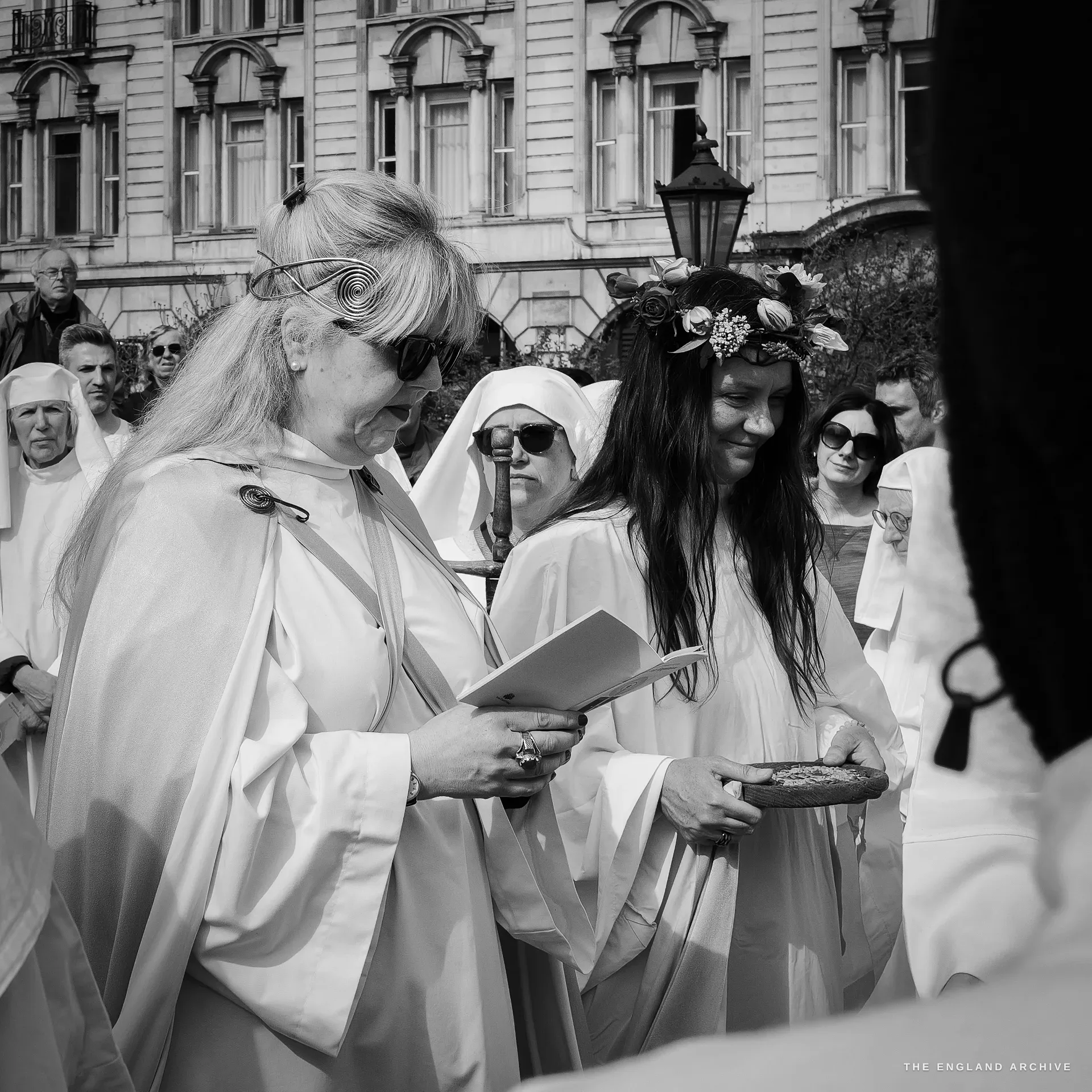 A woman in a white ceremonial robe with a thick floral wreath crowning long dark hair, holding a flat round oat cake in both hands, other Order members around her, a brick London building behind.