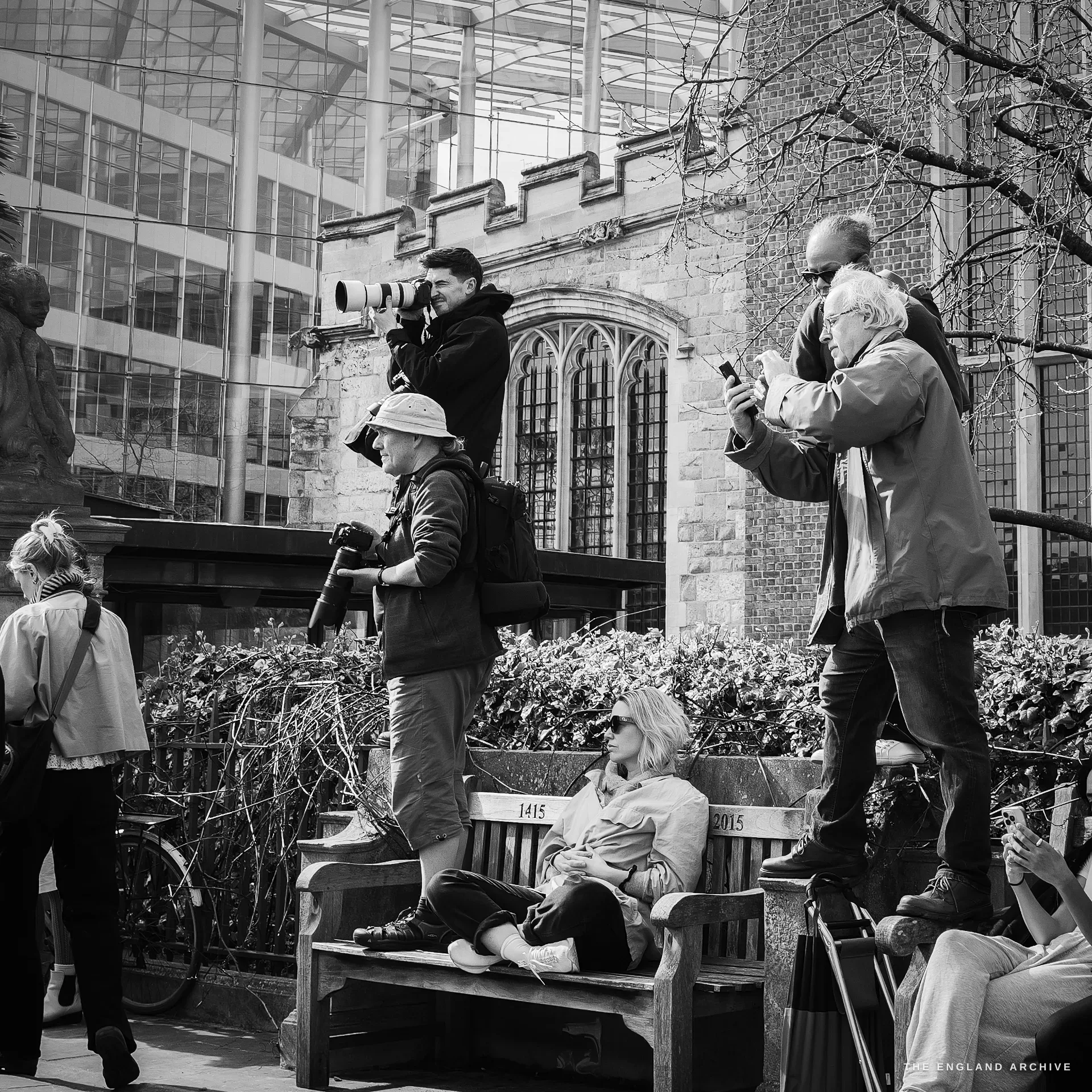 Three press photographers standing on a low wooden bench on Tower Hill, two with long telephoto lenses raised, one shooting on a phone, the gothic stained-glass window of a stone building behind them.