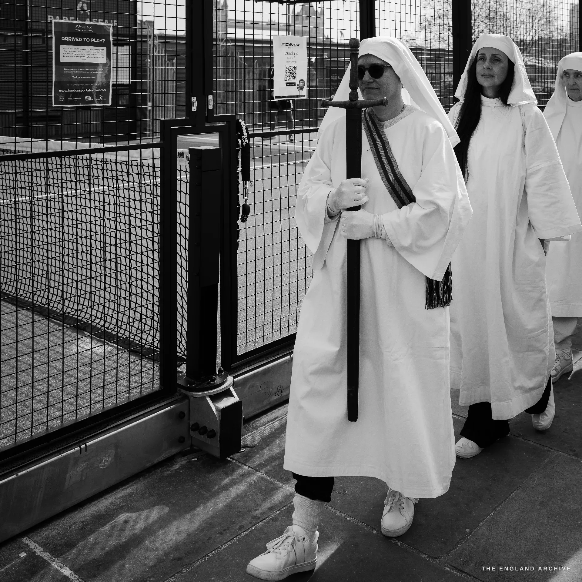 A senior Druid in white robes with sunglasses and a striped scarf carrying a tall vertical wooden staff with a hooked top, walking past a black metal padel-court fence, two other Order members following him.
