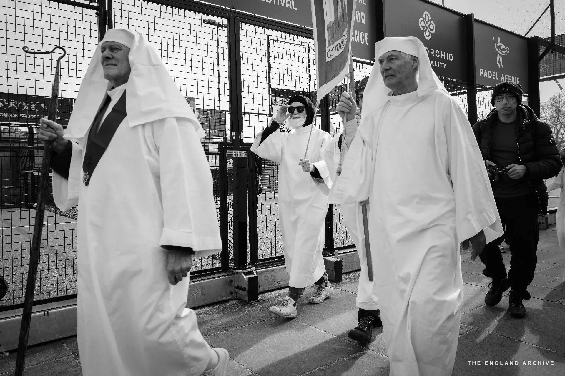 Two senior male Druids walking close in white robes and head veils, padel-court mesh fence behind them, two other Order members trailing in the middle distance.