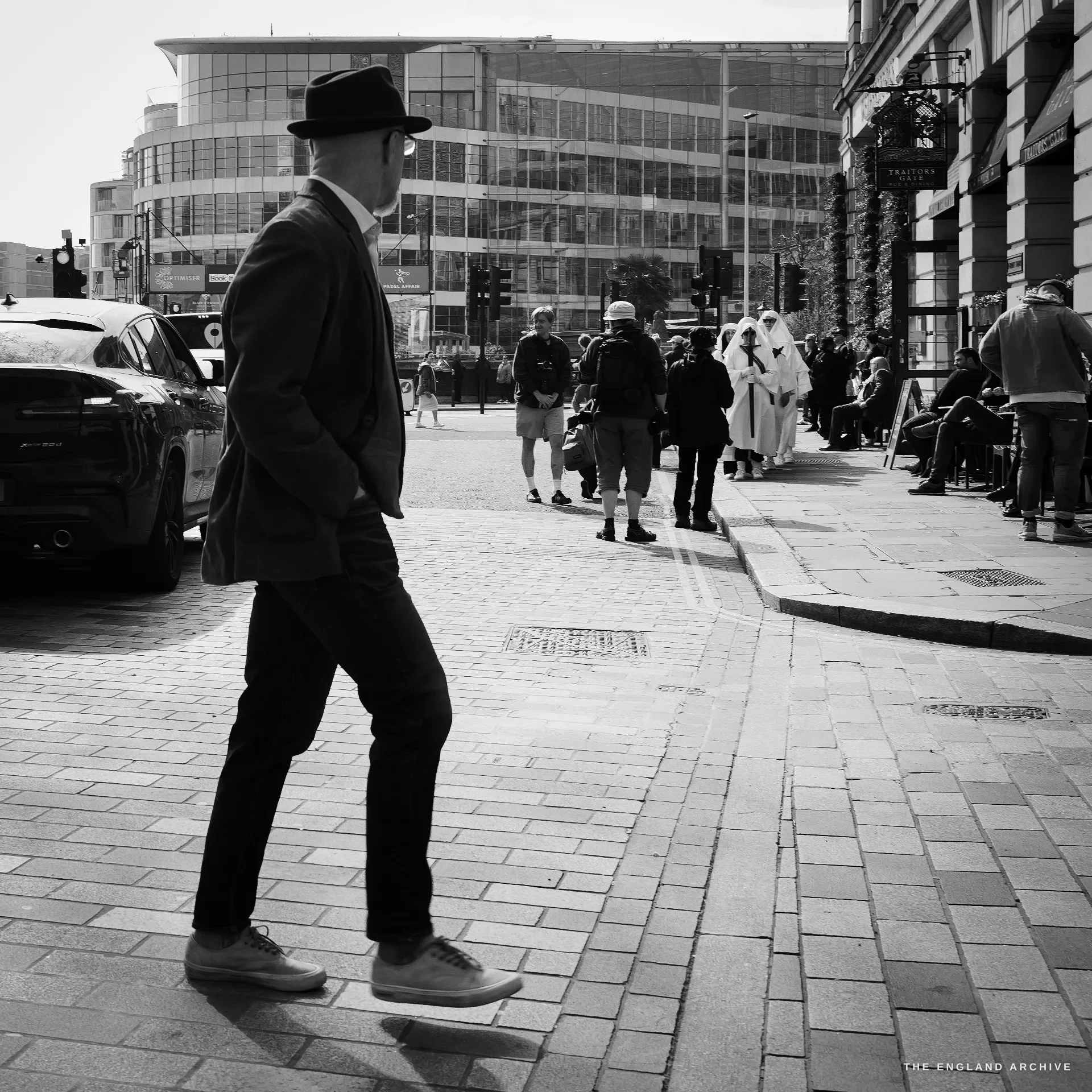 A man in a dark wool coat and brimmed black hat walking briskly across a paved City crossing, sunlight glancing on the pavement; in the middle distance behind him a small group of Druid Order members in white robes are gathered at the kerb.