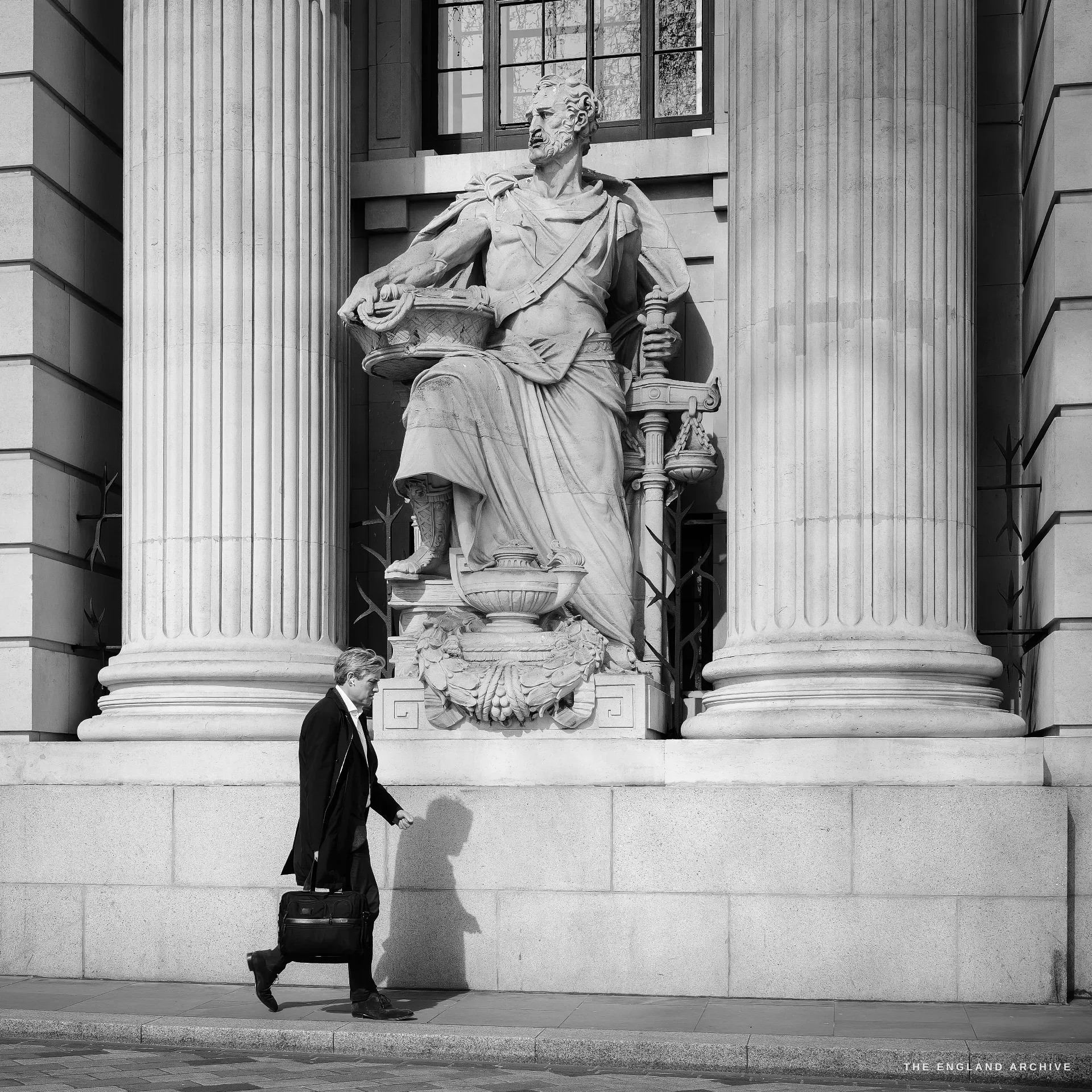 A small businessman walking right-to-left past a colossal classical statue of a seated robed figure mounted on a stone pedestal between fluted columns, the architecture of the Port of London Authority building rising above him.