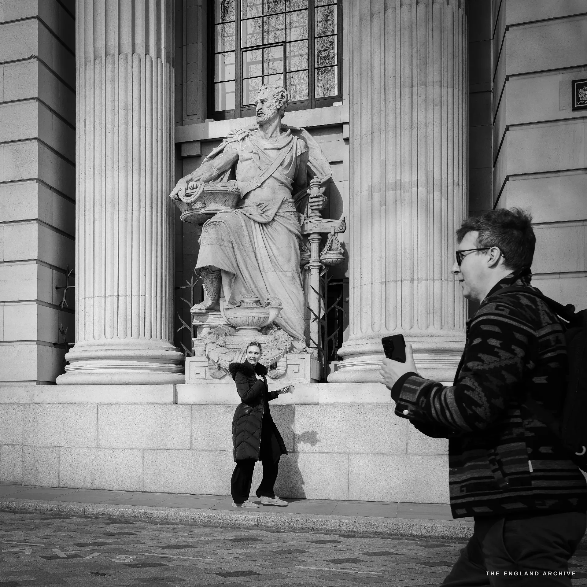 A young woman in a black coat raising a phone to take a selfie at the foot of the Port of London Authority statue, a male photographer in a knit cardigan in the foreground right also raising a phone.