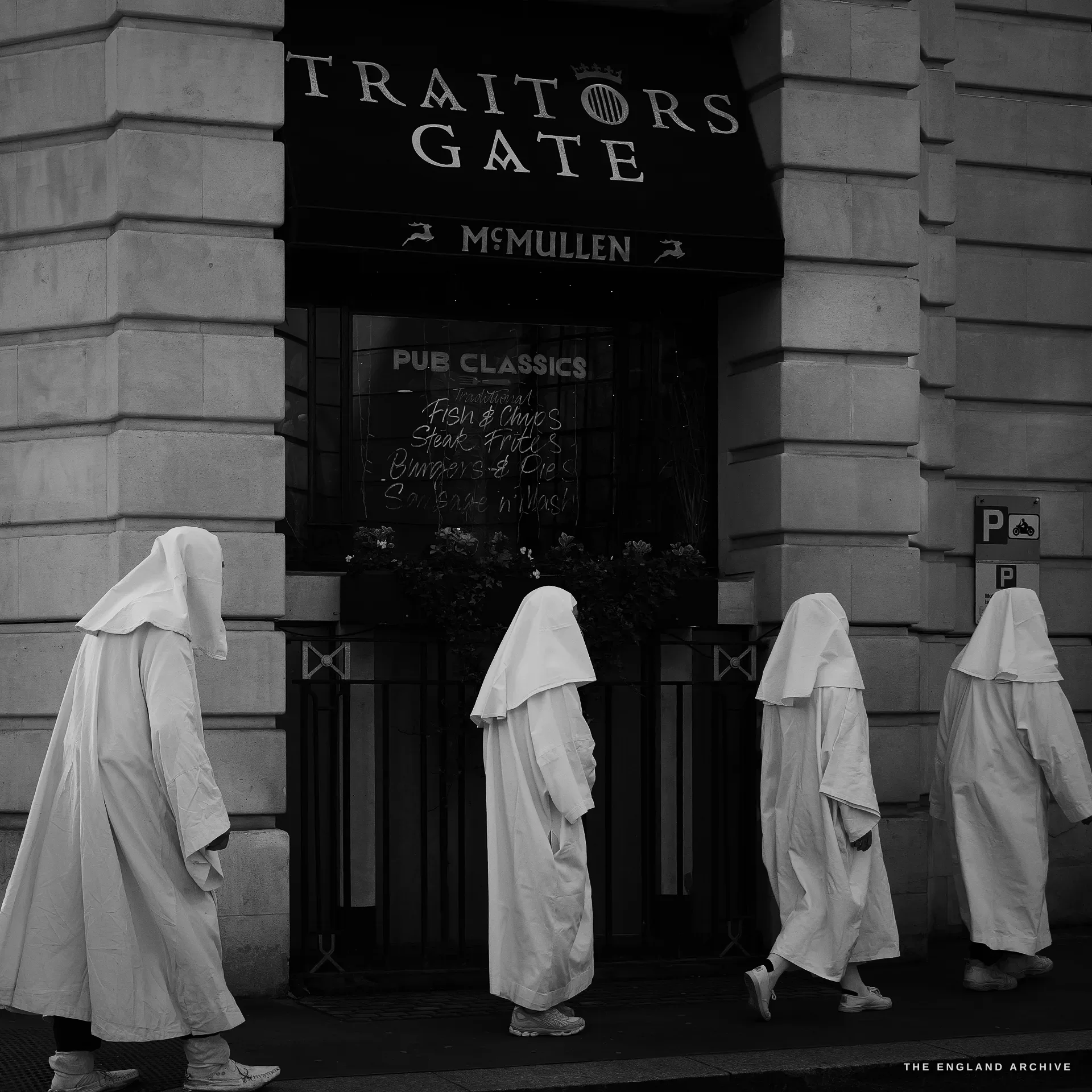 Four Druid Order members in white robes and head veils walking left-to-right past a stone pub front, the painted awning above them reading TRAITORS GATE / McMULLEN with a chalkboard inside the doorway listing PUB CLASSICS - fish and chips, steak frites, burger and fries.