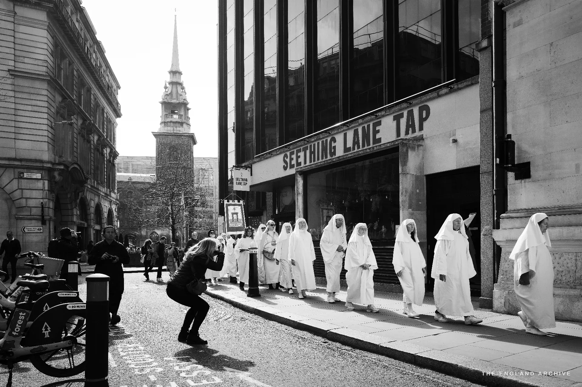 A documentary photographer crouched in the middle of an empty City road photographing a procession of Druid Order members in white robes walking left-to-right along the pavement past a SEETHING LANE TAP shopfront, the brick body and white spire of All Hallows-by-the-Tower visible in the distance.