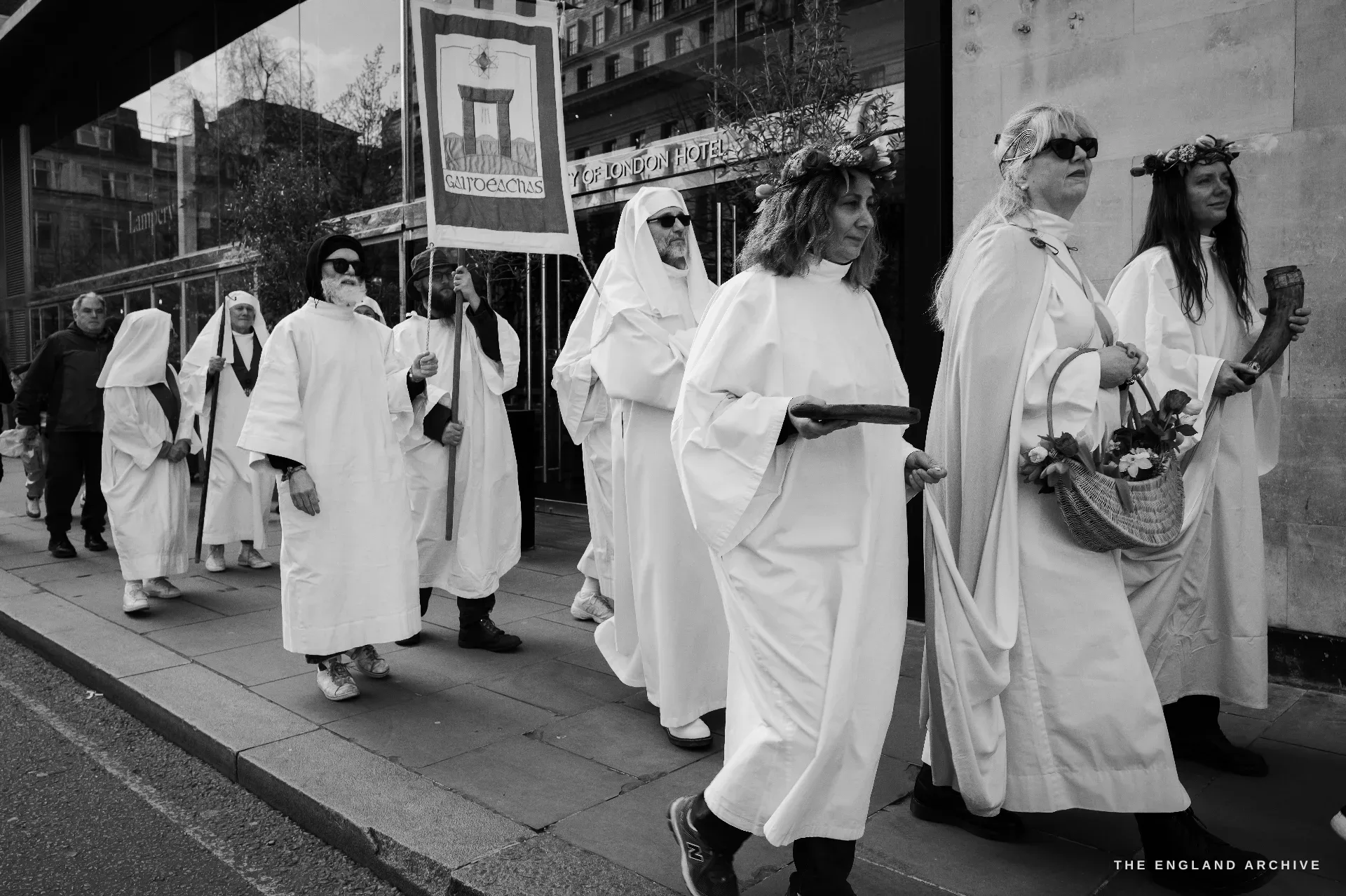 A close procession of Druid Order members in white robes walking left-to-right along a pavement, a banner with an embroidered tower image at the centre, two women at the head of the line in floral crowns, one carrying a small flower basket.