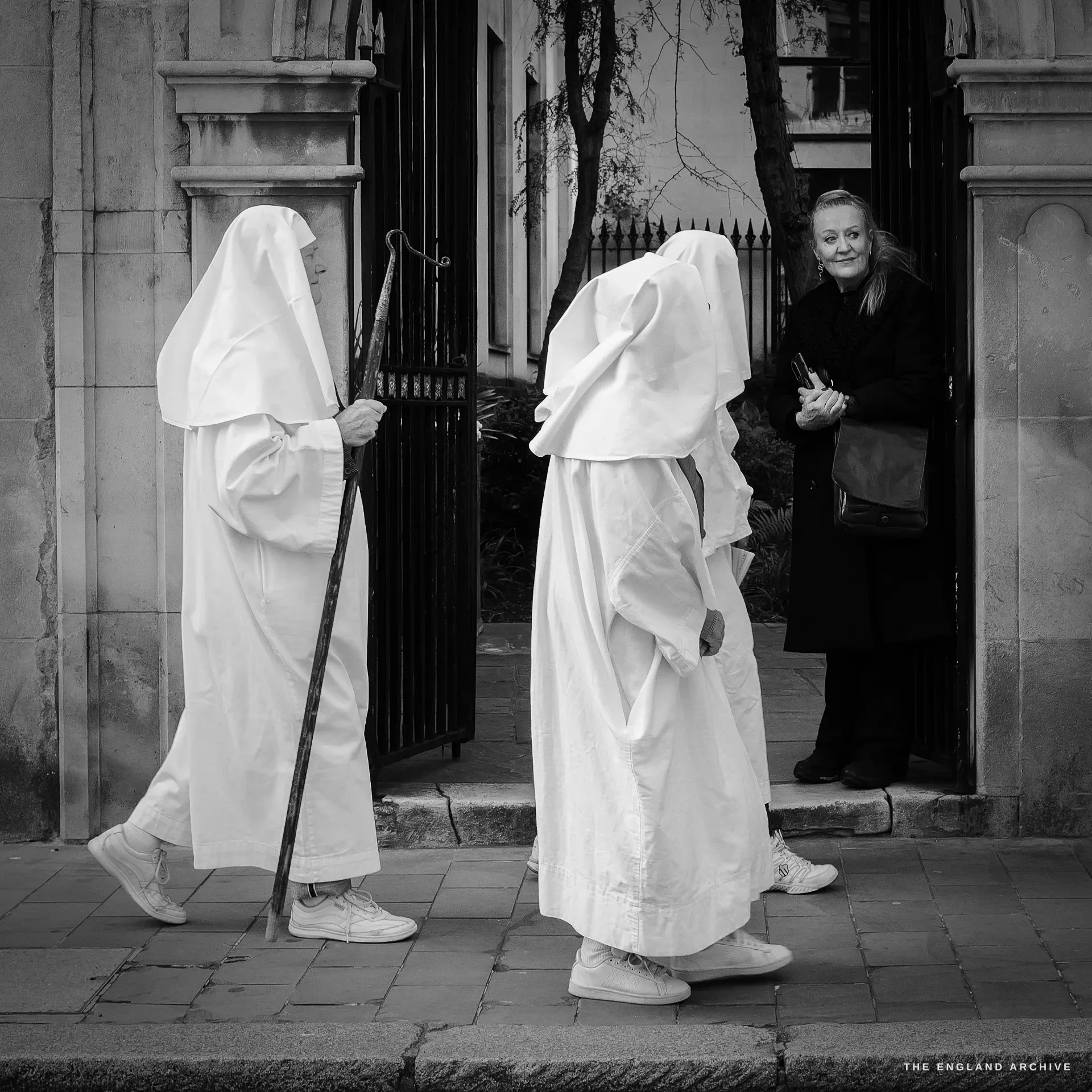 Two robed Druid Order members crossing through a black wrought-iron gate, the leading figure holding a tall hooked wooden staff, a woman in a dark coat watching from the doorway behind to the right.