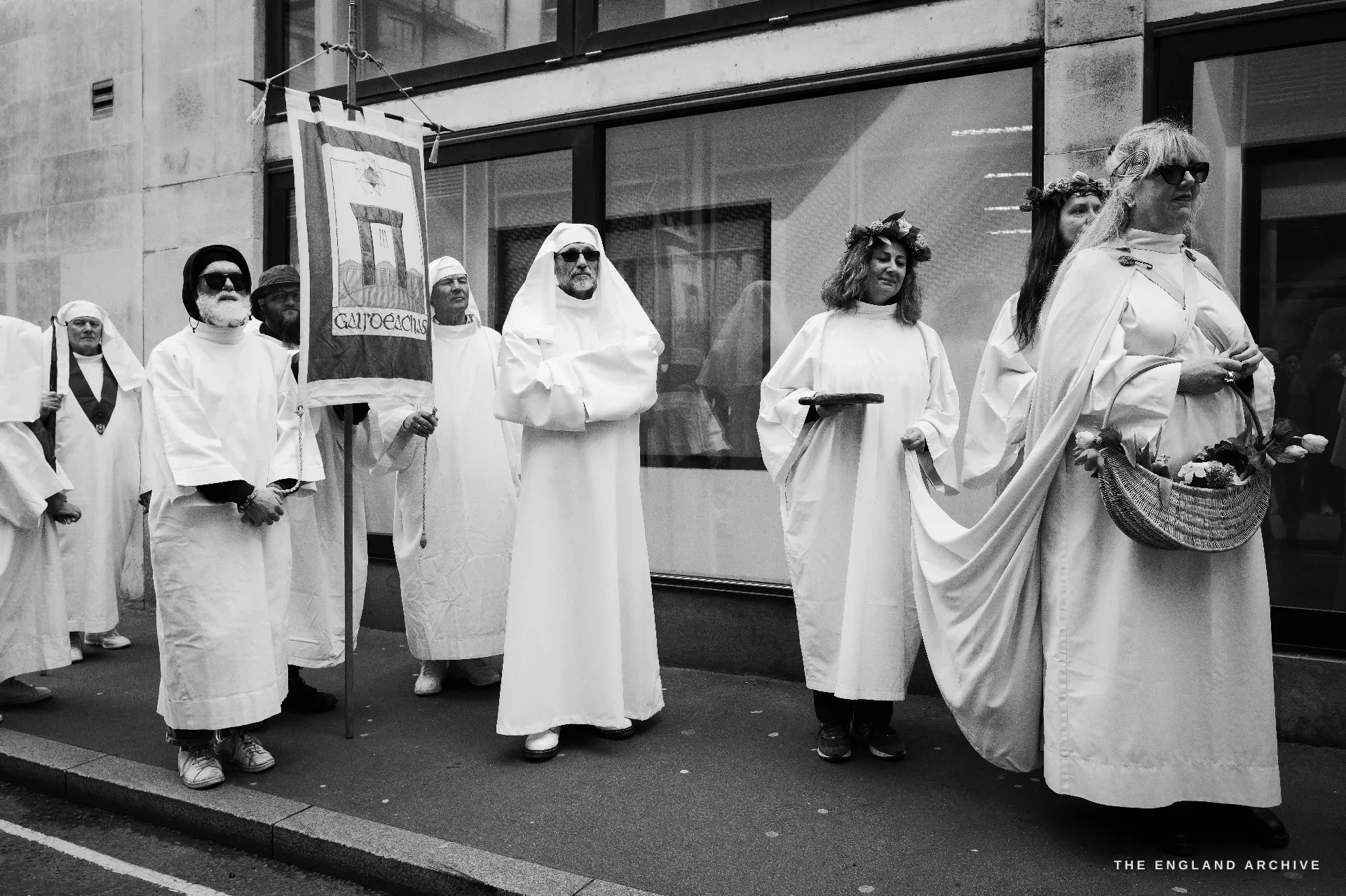 A row of seven Druid Order members standing along a pavement, the embroidered banner of the order at the centre showing a tower and a chalice; one man in dark glasses holds a vertical staff at left, one woman in a floral crown at right.