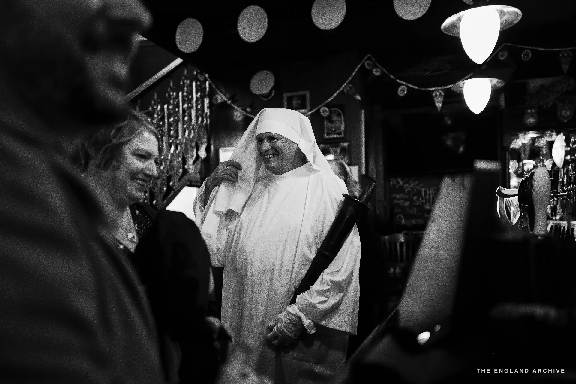 A central Druid in white robes laughing under the warm fairy-light interior of a London pub, a sword carried point-down at his side, fellow members and a publican in conversation around him.