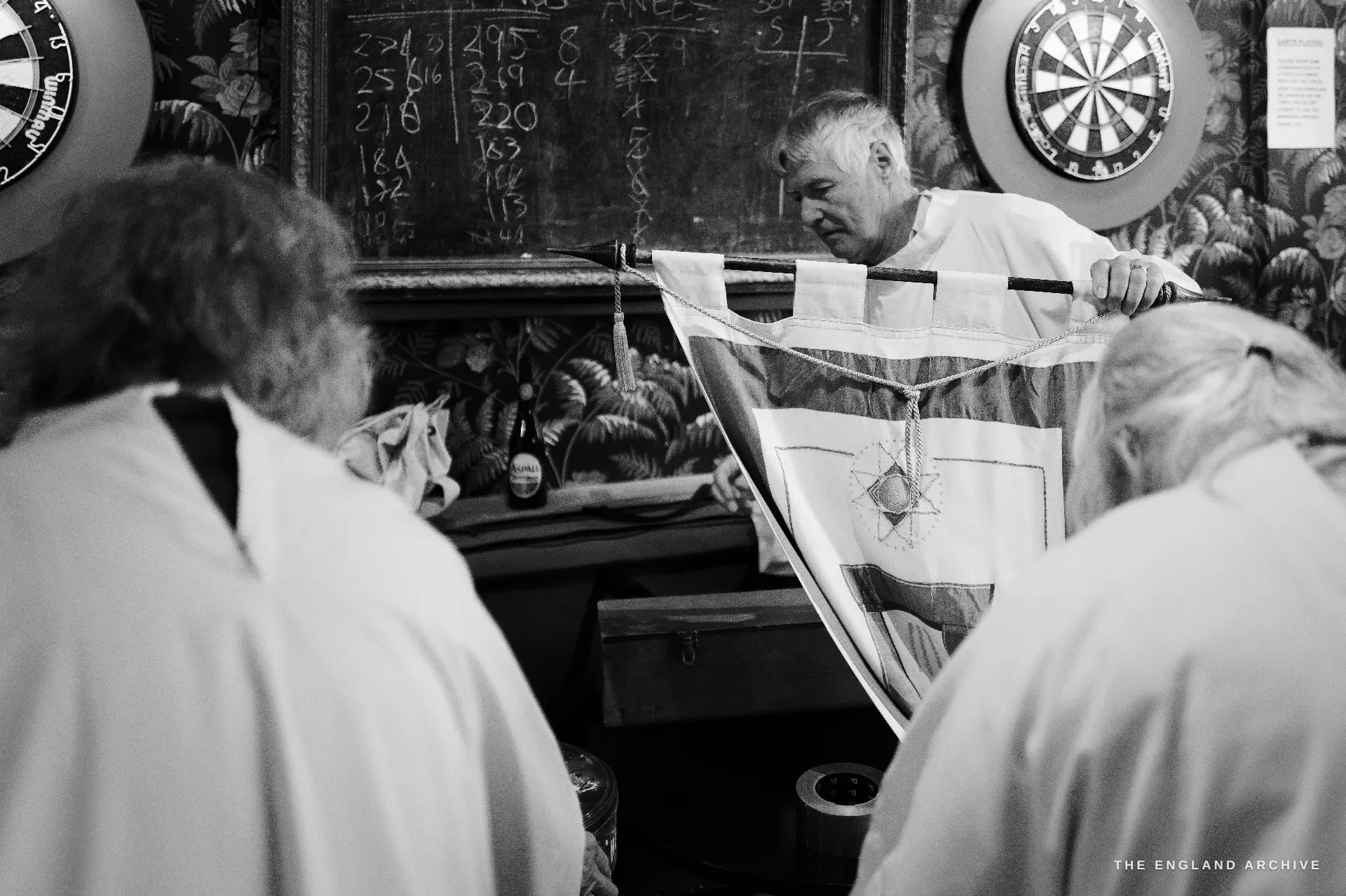 A Druid in white robes holding up a large embroidered ceremonial cloth with a hexagram-and-cross design, two dart boards mounted on the dark-panelled pub wall behind him, a pub chalkboard scoreboard with hand-written numbers visible to the left.