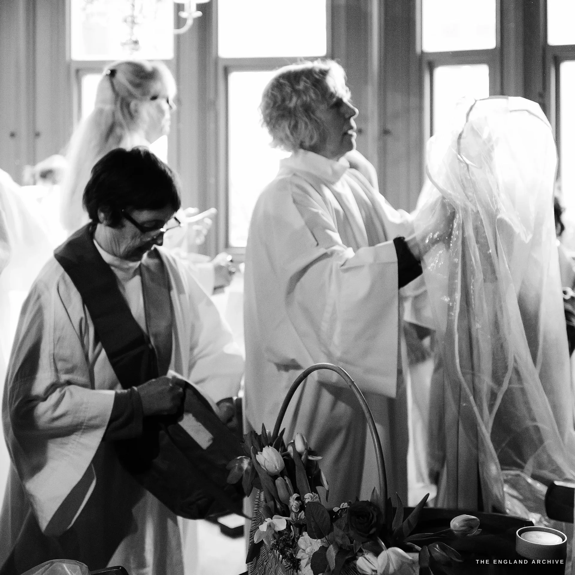 Inside a high-windowed function room, a senior female Druid helps another member adjust a long flowing white veil, both in white robes; in the foreground a wicker basket of flowers, tulips, and ferns rests on a table, a tall pillar candle to the right.