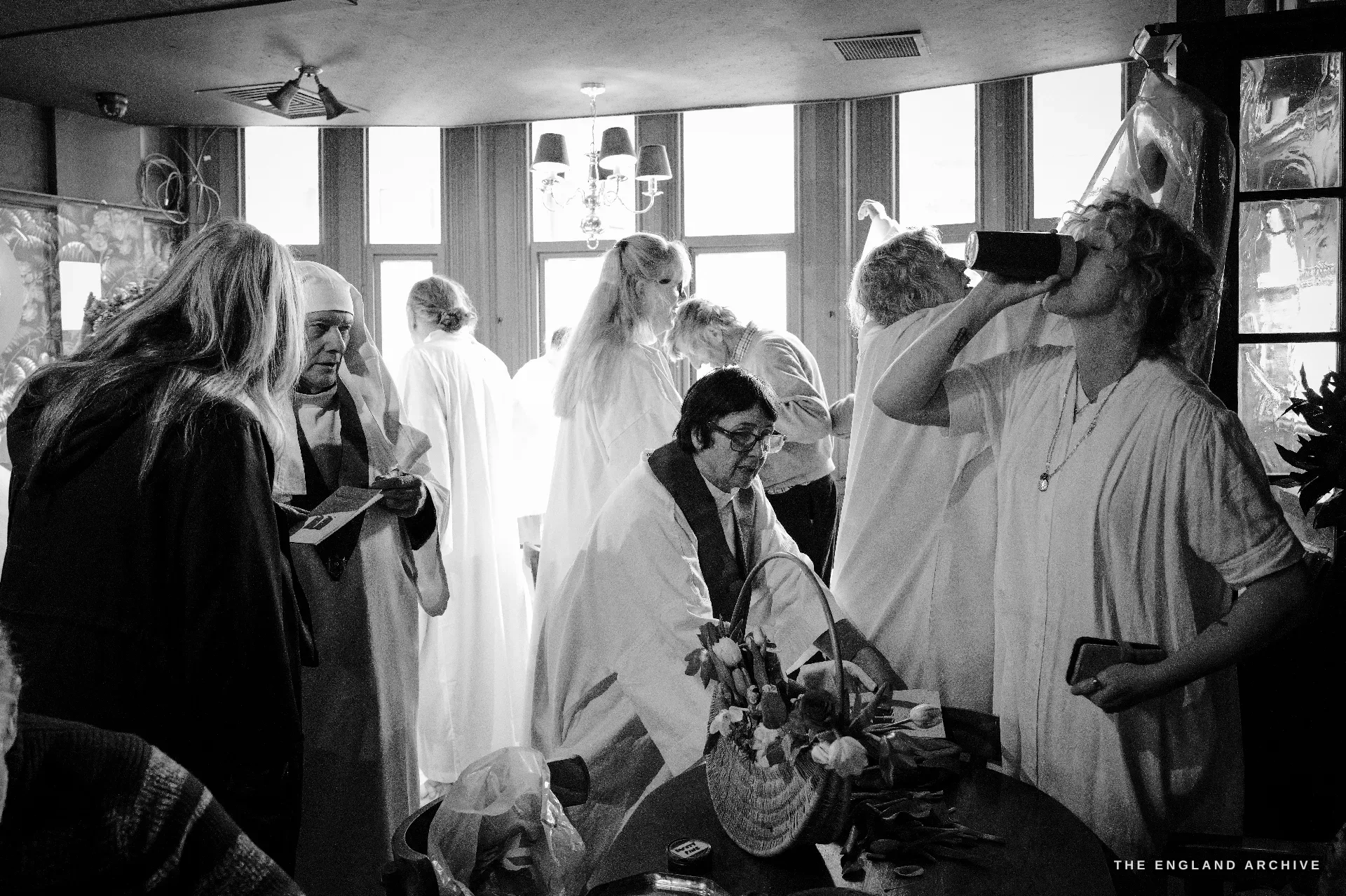 A high-windowed pub function room full of Druid Order members in white robes in casual conversation, a man at the centre lifting a pint glass to his lips, a wicker basket of flowers and a vase of tulips on the table in the foreground.