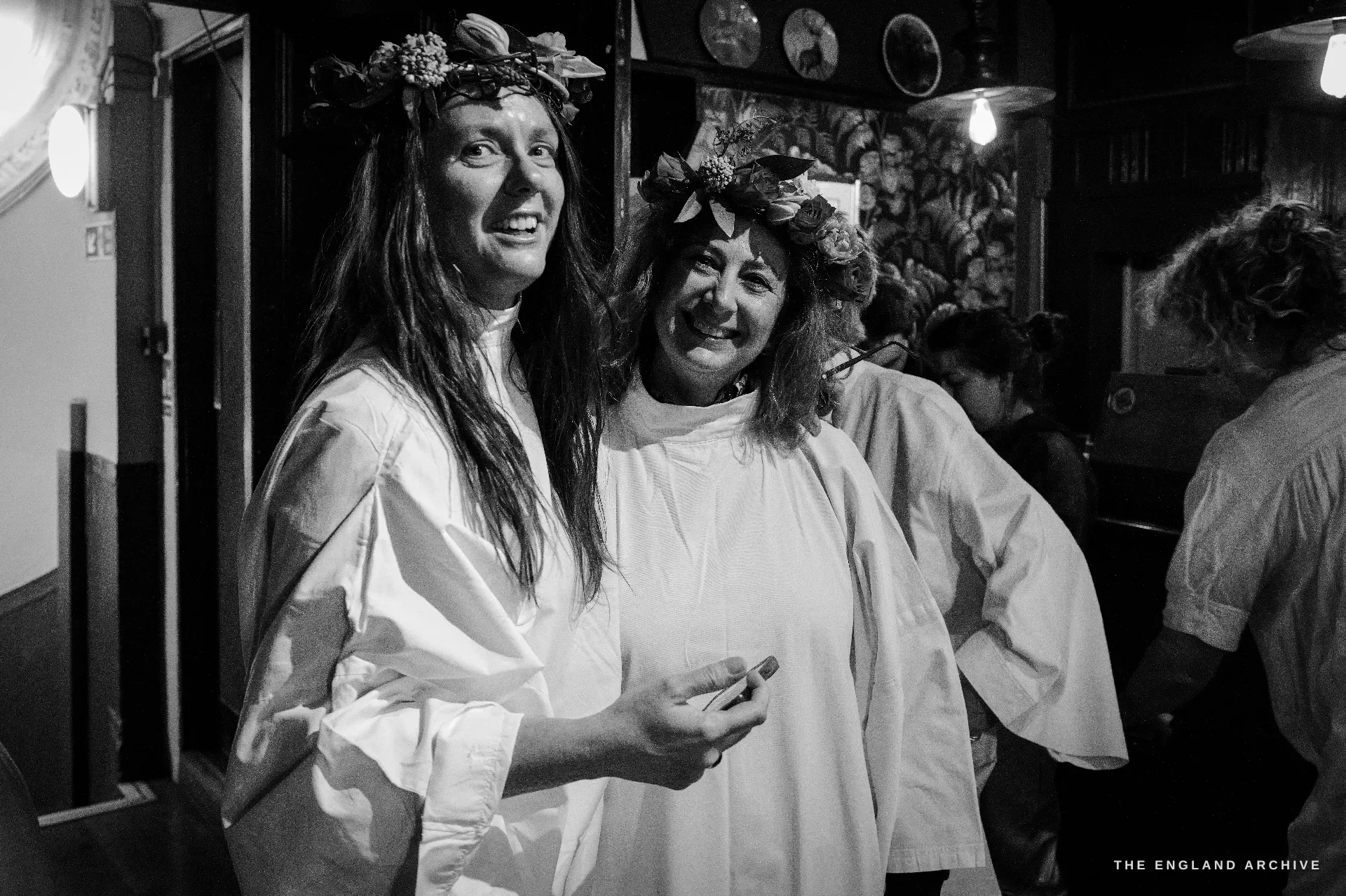 Two female Druid Order members close together in the warm pub interior, both wearing floral crowns, both smiling, one with long brown hair, the other with a small flower wreath, fairy lights behind them.