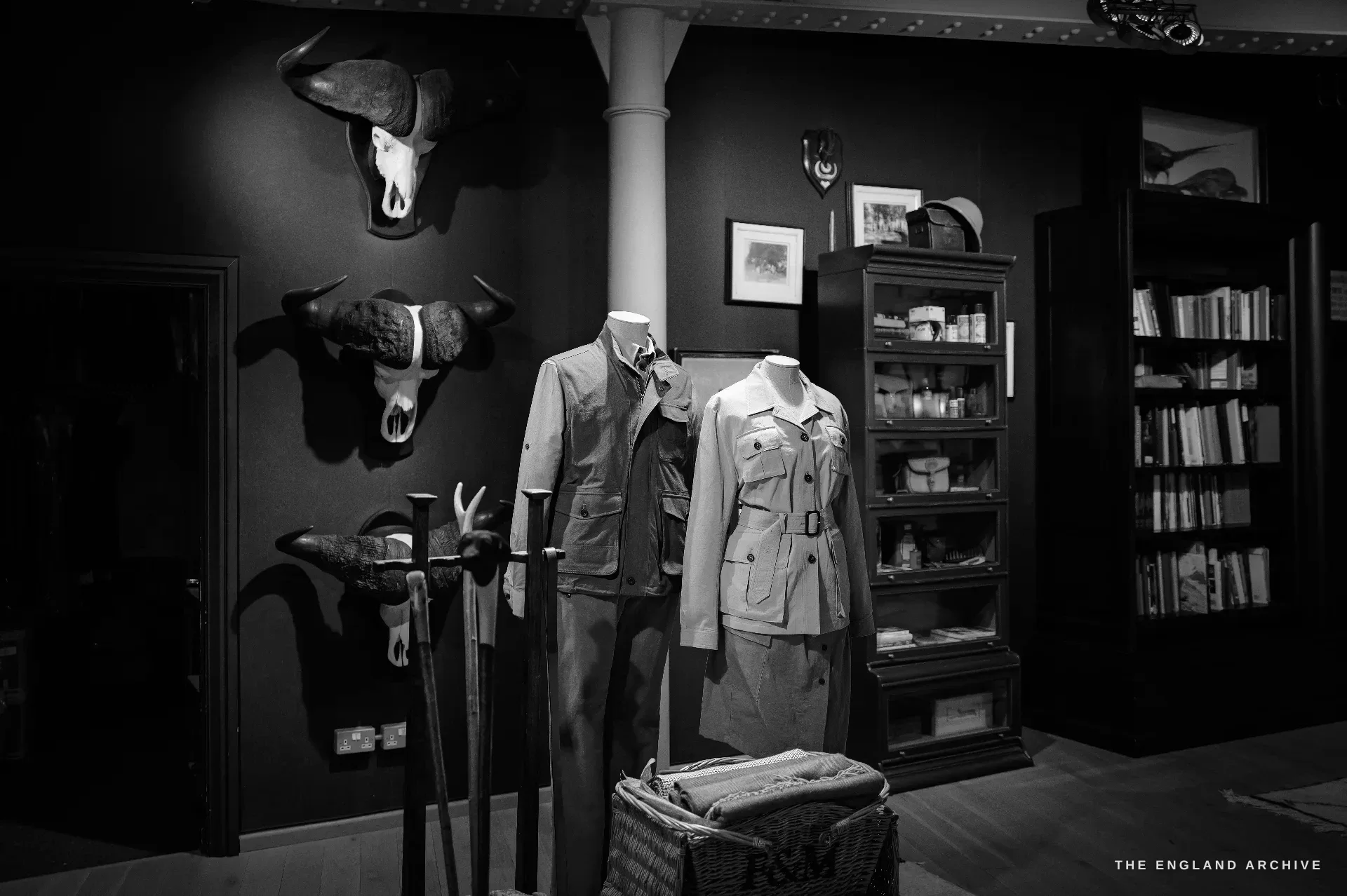 A showroom wall hung with three cape buffalo skull mounts above two mannequins in safari clothing, a wicker Fortnum & Mason hamper, a bookcase and shooting sticks.