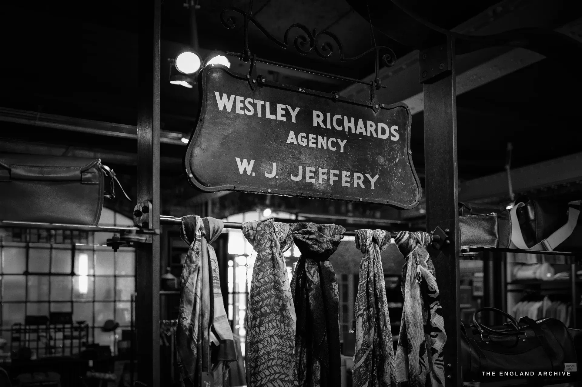 A hanging wrought-iron sign with gilt lettering reading WESTLEY RICHARDS AGENCY, W. J. JEFFERY, above a rail of patterned silk scarves and leather bags.