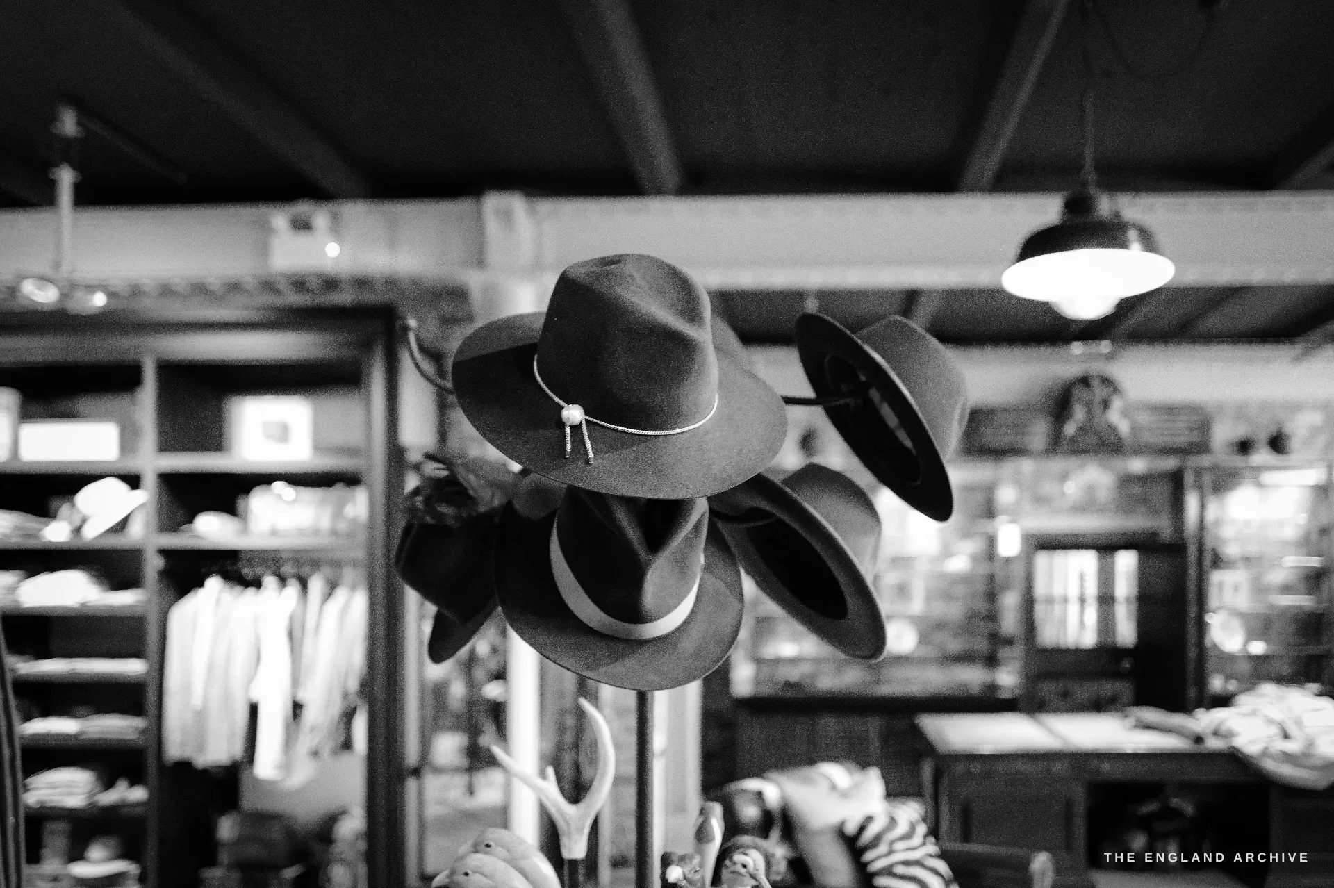 A stand of felt sporting hats in the outfitting room, shelves of folded clothing and glazed cabinets softly lit behind, a pendant lamp overhead.