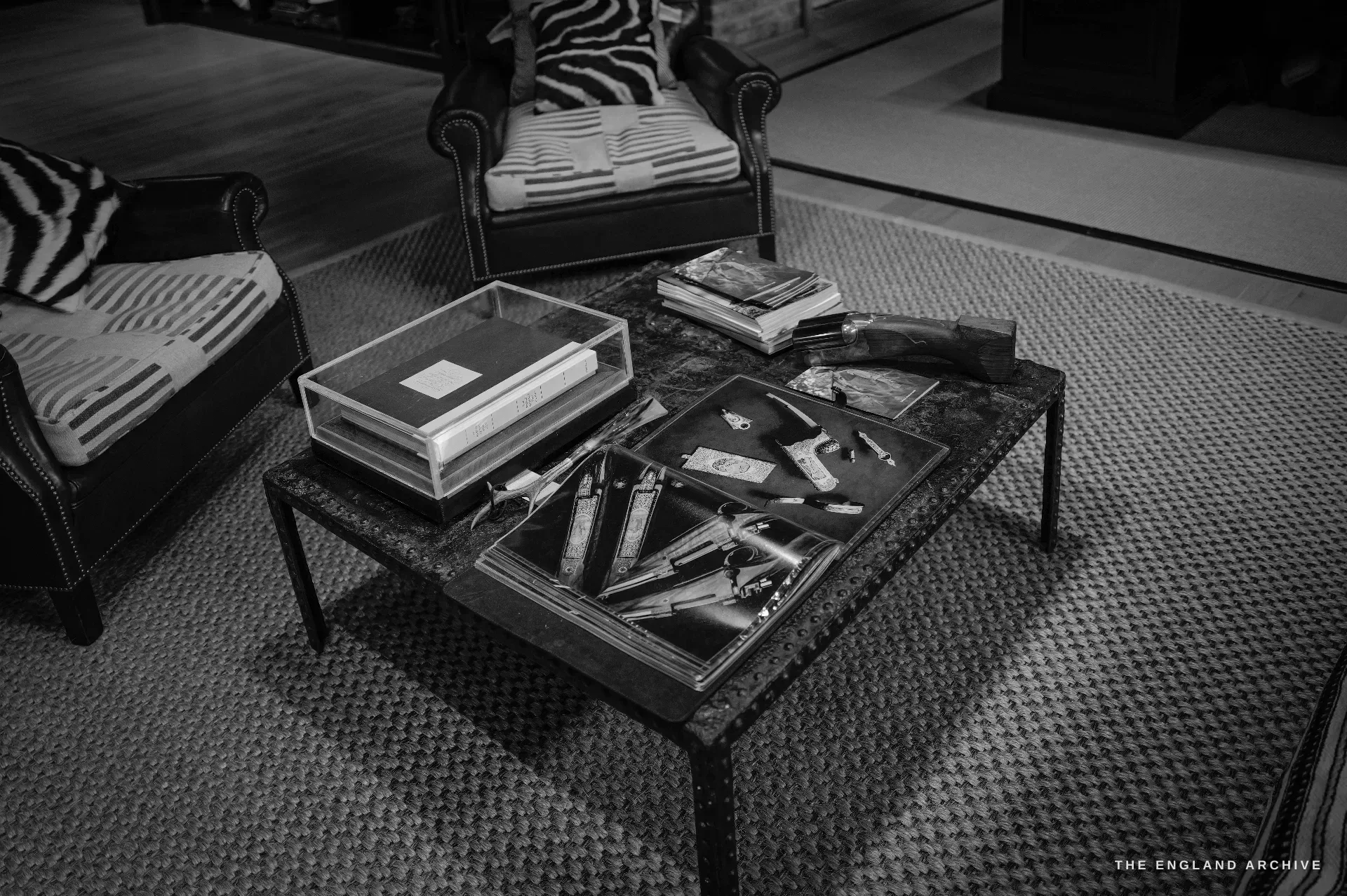 A low iron coffee table between leather armchairs, holding a cased record book, stacks of catalogues, and a large open book showing photographs of engraved gun actions, a shotgun laid across the table.