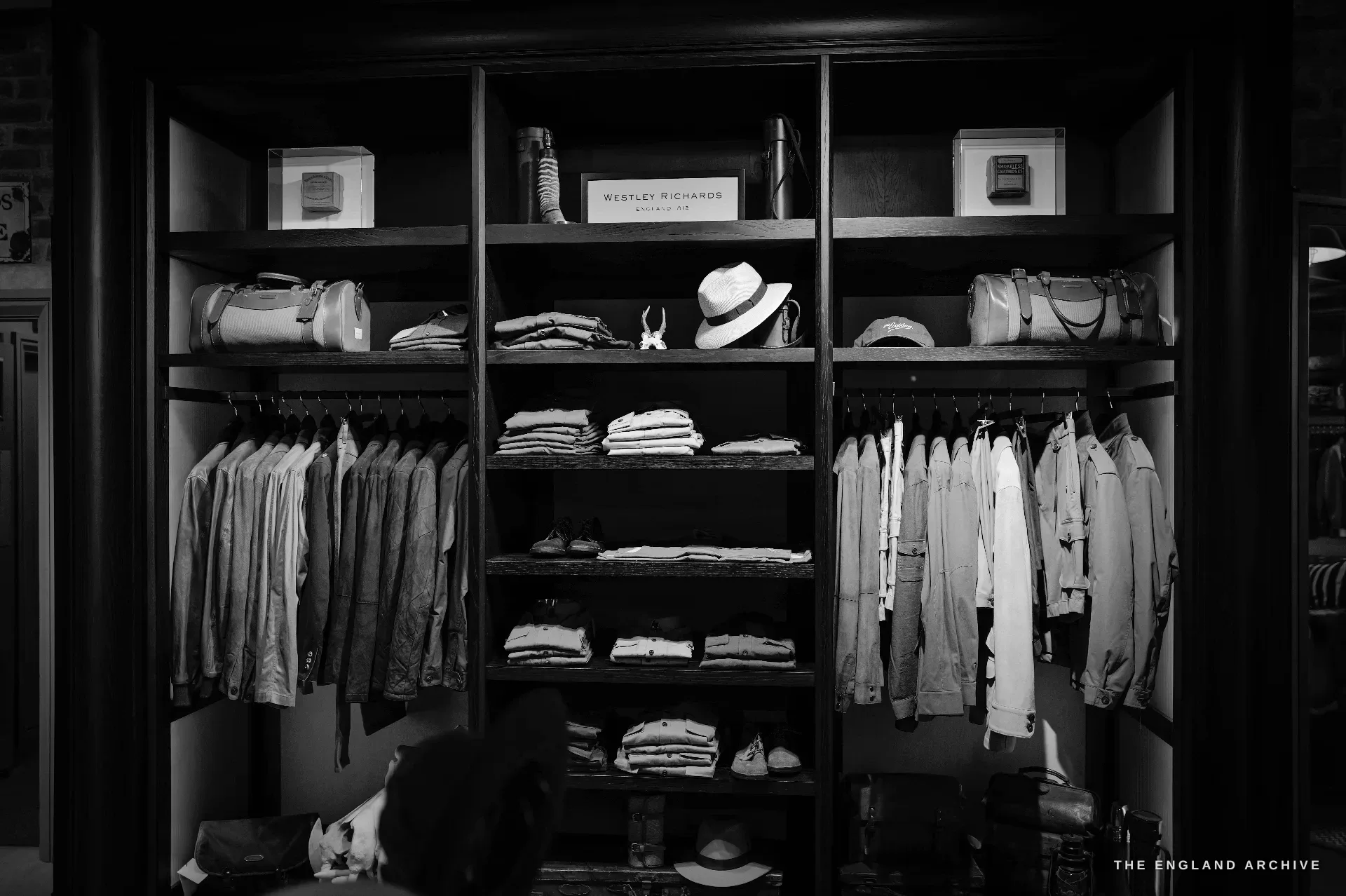 A dark display shelf of folded tweed and cotton clothing, a panama hat, leather bags and a printed card reading WESTLEY RICHARDS, ENGLAND 1812.