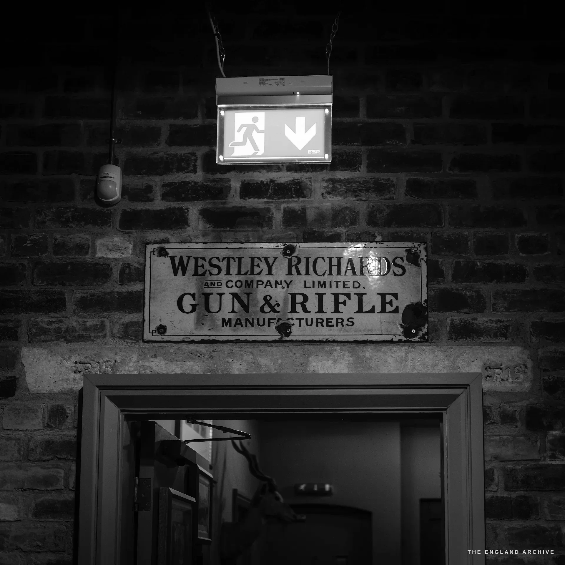 A weathered enamel sign fixed to a brick wall above a doorway reading WESTLEY RICHARDS AND COMPANY LIMITED, GUN & RIFLE MANUFACTURERS, a green emergency-exit sign glowing above it.