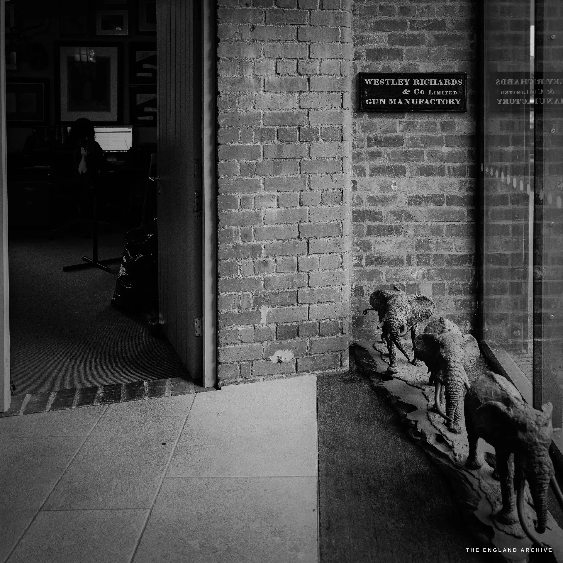The manufactory entrance seen from inside the glass doors: a cast plaque on the brick reading WESTLEY RICHARDS & Co Limited GUN MANUFACTORY, a cast-iron elephant boot-scraper on the mat, and the car park beyond.